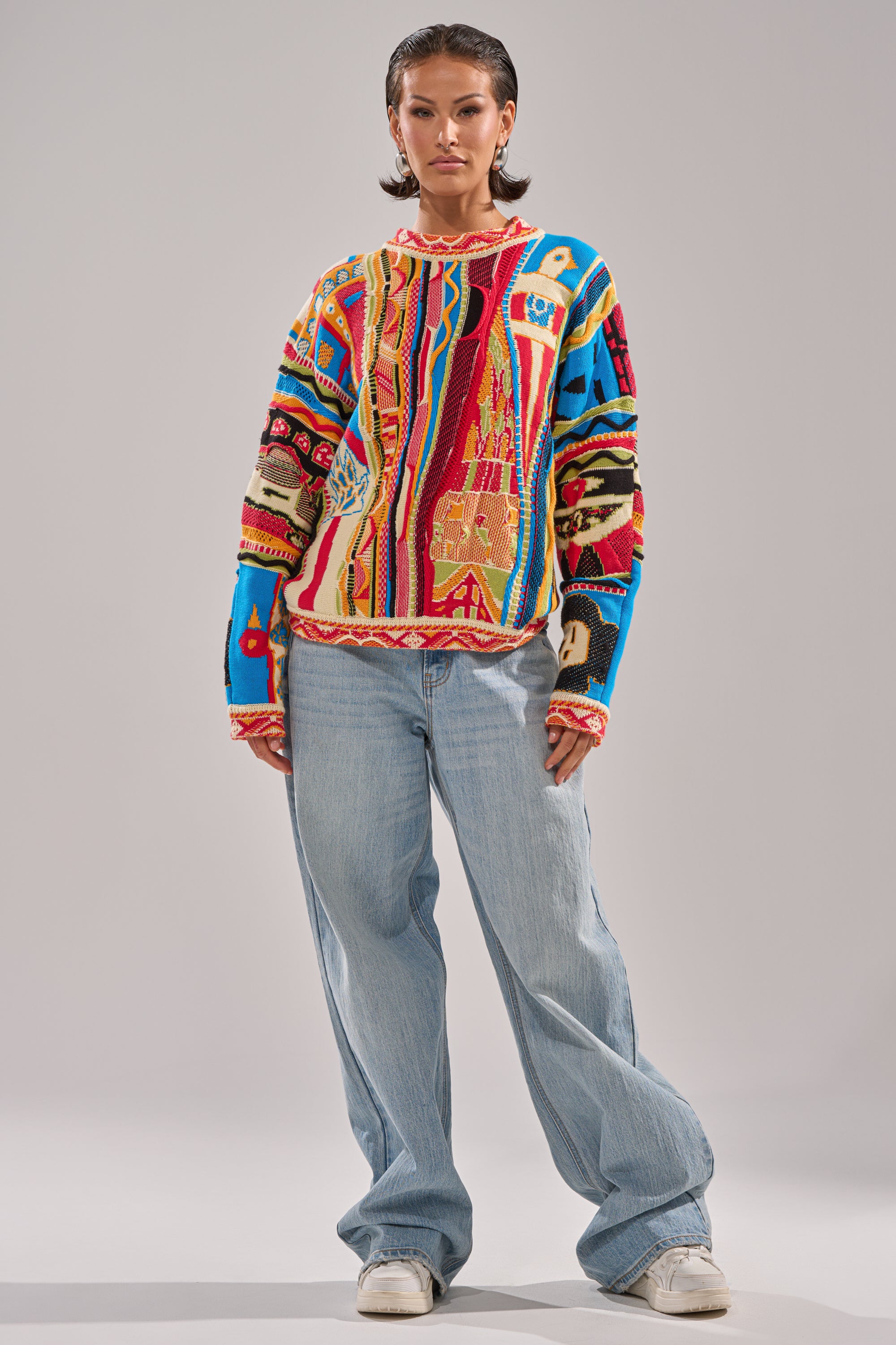 A person with short dark hair and a neutral expression stands against a plain backdrop, wearing the RETRO OVERSIZED SWEATER IN MULTI, loose light blue jeans, and white sneakers.