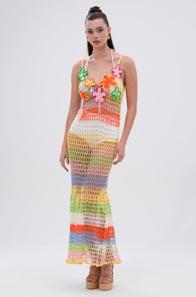 Wearing the BEACHIN IT CROCHET BEADED MAXI DRESS, a woman poses against a white background. The dress features vibrant stripes and large floral appliqués, is sleeveless and sheer, and is styled with platform sandals.