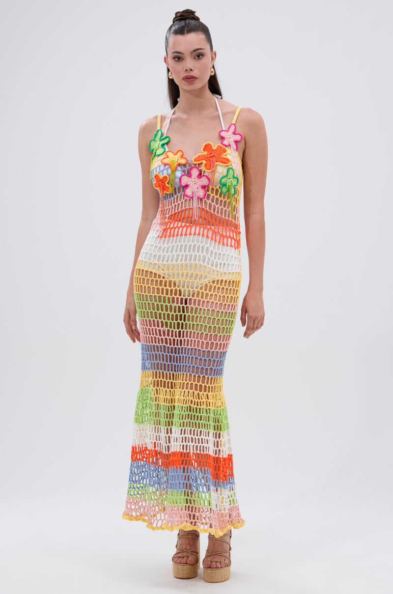 A woman with long, straight hair pulled back faces forward in the BEACHIN IT CROCHET BEADED MAXI DRESS, a colorful ankle-length dress featuring large multicolored floral appliqués and a sheer netted design. She pairs it with platform sandals.