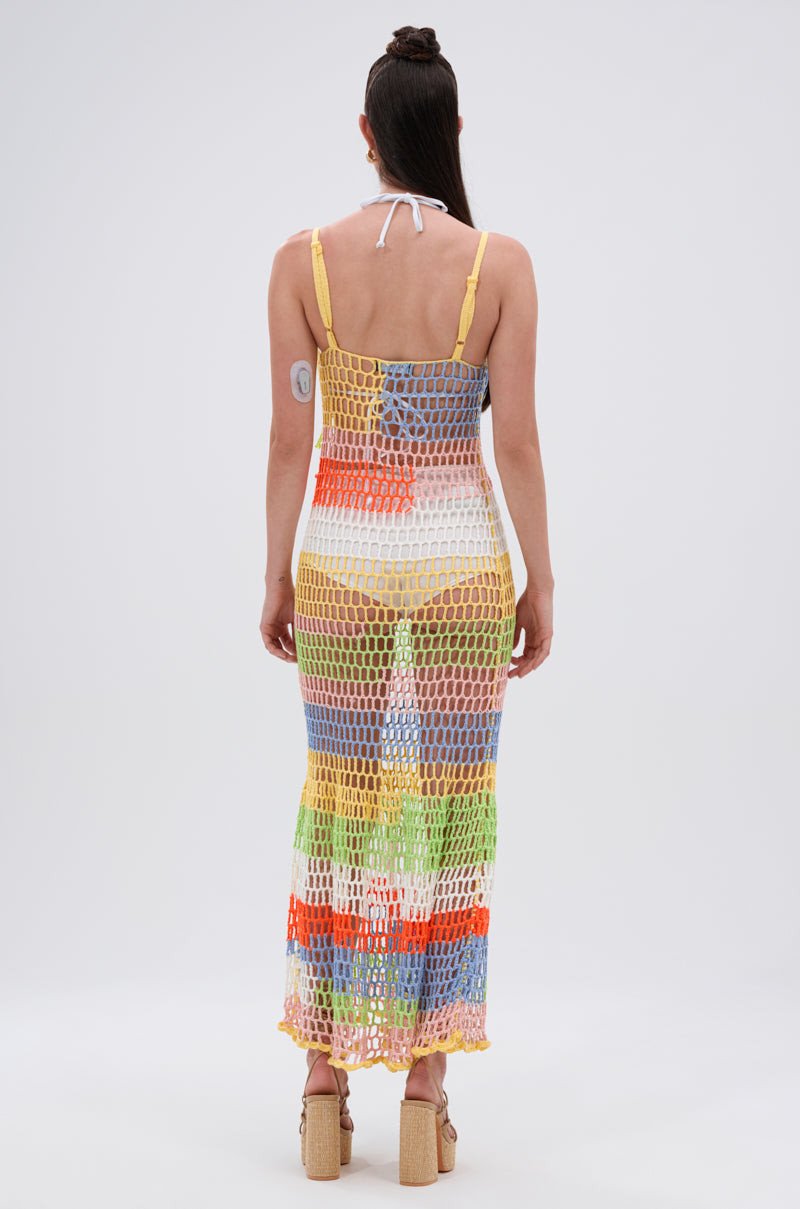 A woman with long brown hair and beige platform sandals faces away, modeling the BEACHIN IT CROCHET BEADED MAXI DRESS with large holes over a swimsuit. An arm patch is visible. The background is plain white.