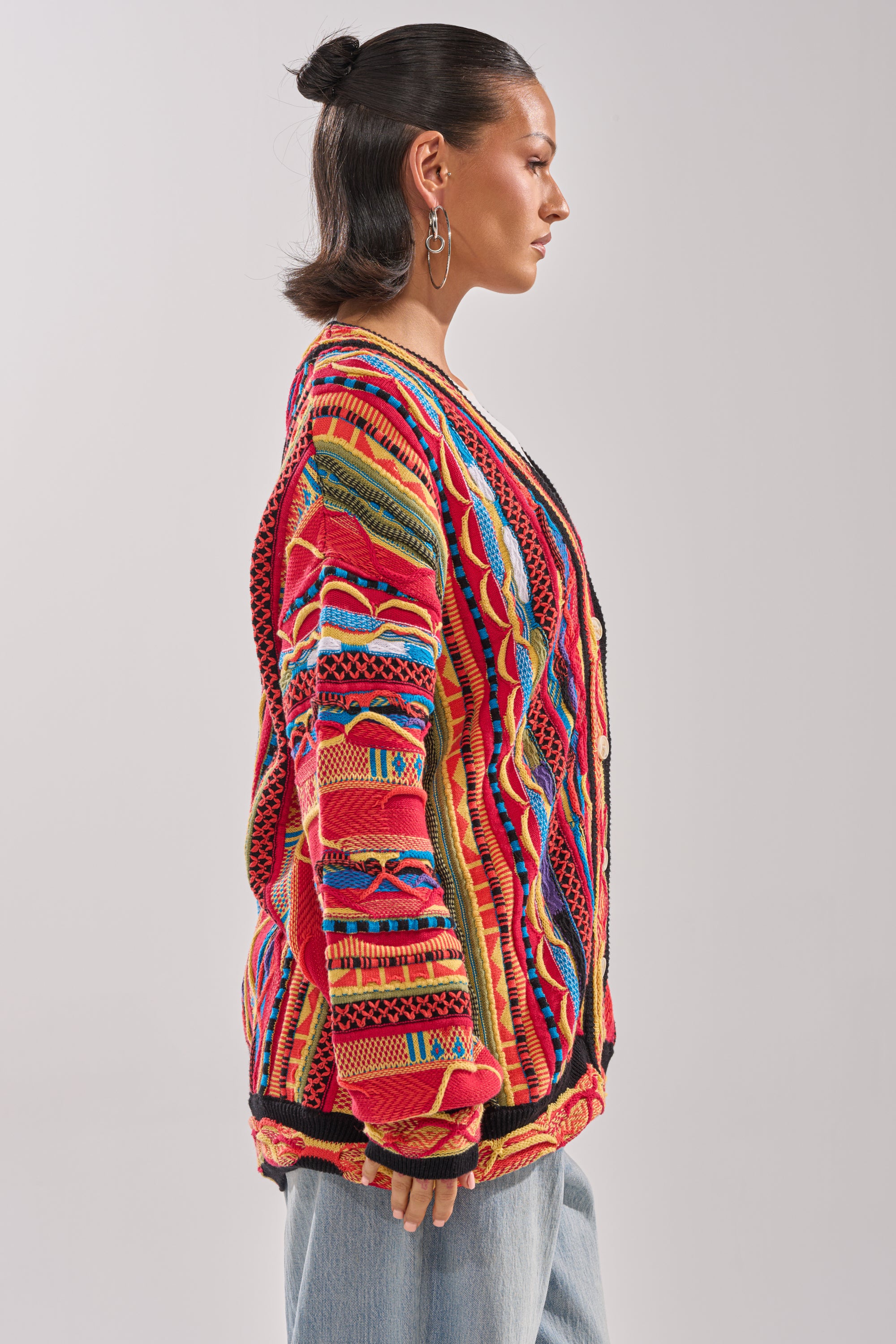 A person stands in profile wearing the CRAZY IN LOVE CARDIGAN, a retro knit sweater with vibrant red, yellow, blue, and green patterns, paired with light blue jeans against a plain light gray background. 3