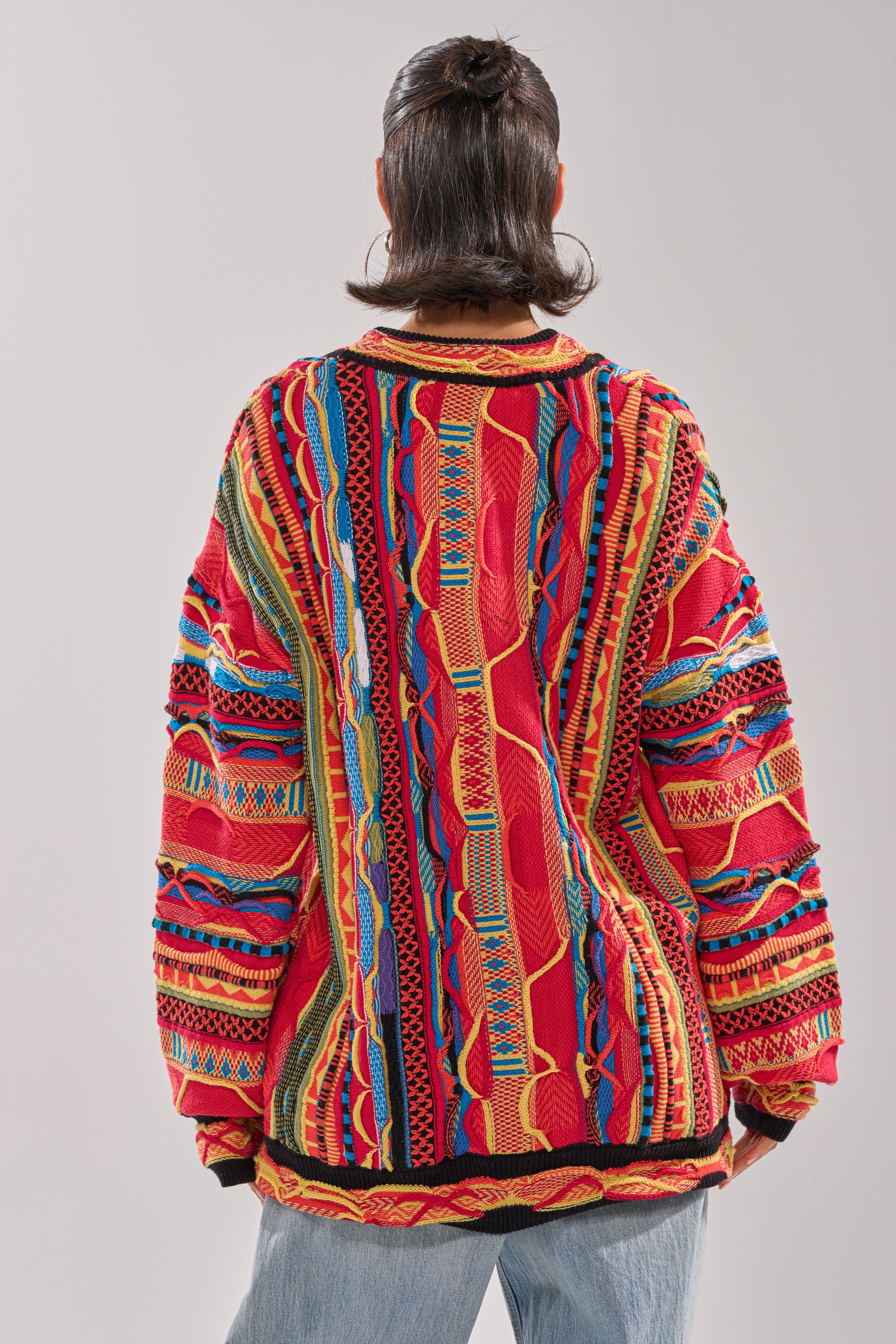 A person stands with their back turned, wearing the CRAZY IN LOVE CARDIGAN— a colorful retro knit featuring bold red, yellow, blue, and green geometric patterns— paired with light blue jeans and shoulder-length hair. 4