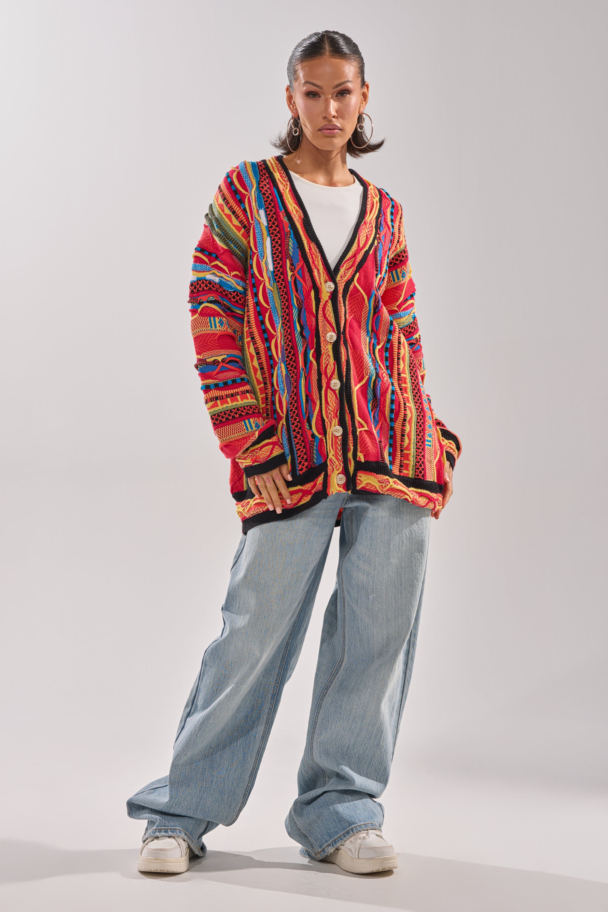 A woman wears the CRAZY IN LOVE CARDIGAN, a colorful retro knit, over a white shirt with wide-leg light blue jeans and white sneakers. She accessorizes with hoop earrings and has two ponytails, set against a plain light background. 5