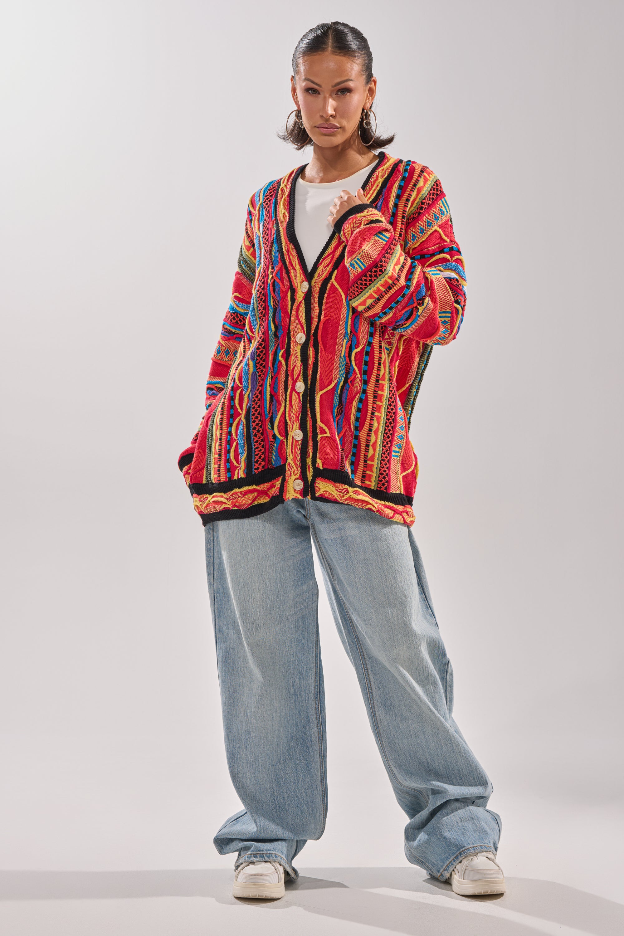 A person stands confidently in the CRAZY IN LOVE CARDIGAN, a colorful retro knit sweater, paired with a white top, wide-leg light blue jeans, and white sneakers against a plain light background. 6