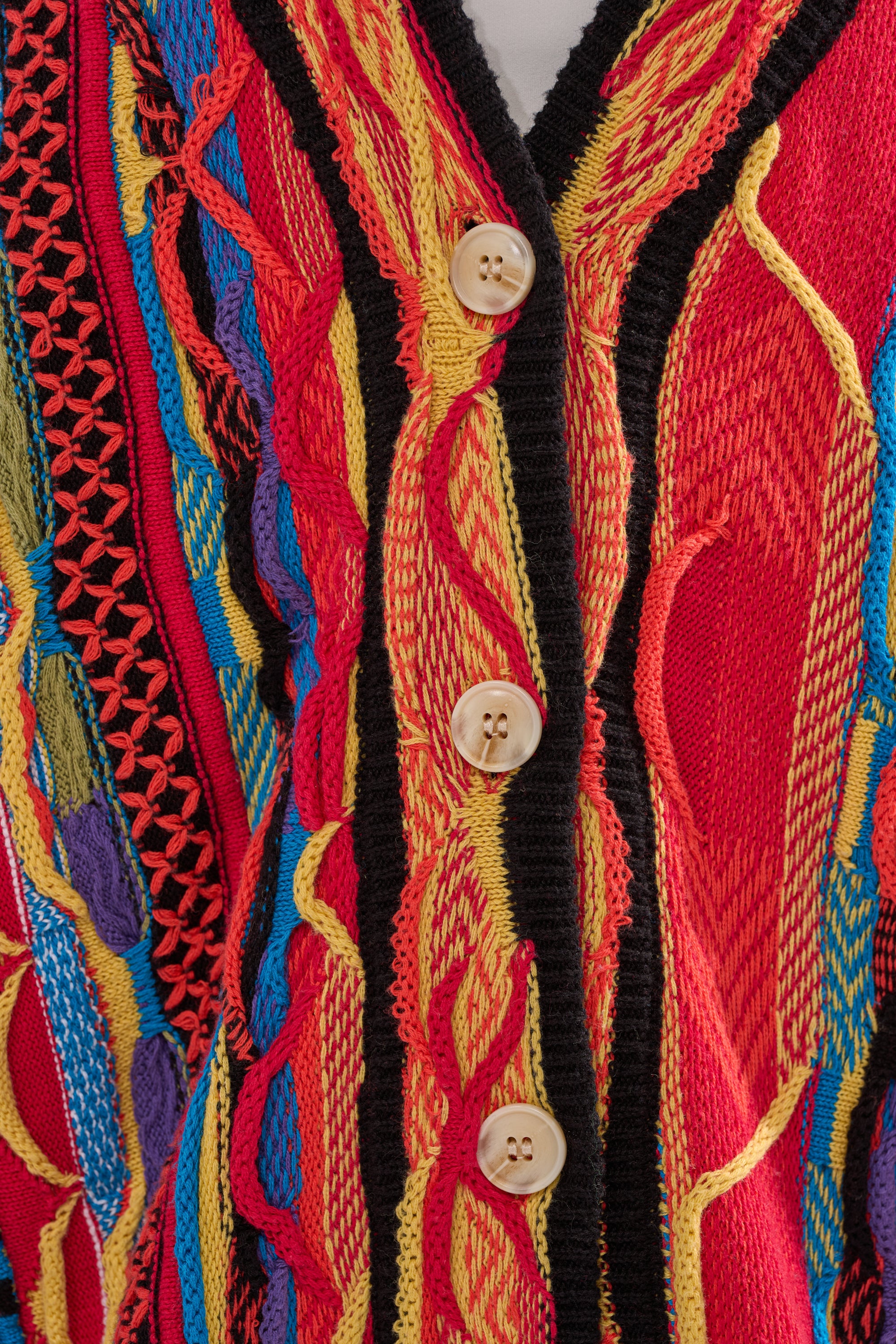 Close-up of the CRAZY IN LOVE CARDIGAN, a colorful retro knit sweater with red, yellow, blue, and purple patterns and three tan buttons down the black-trimmed front. 7