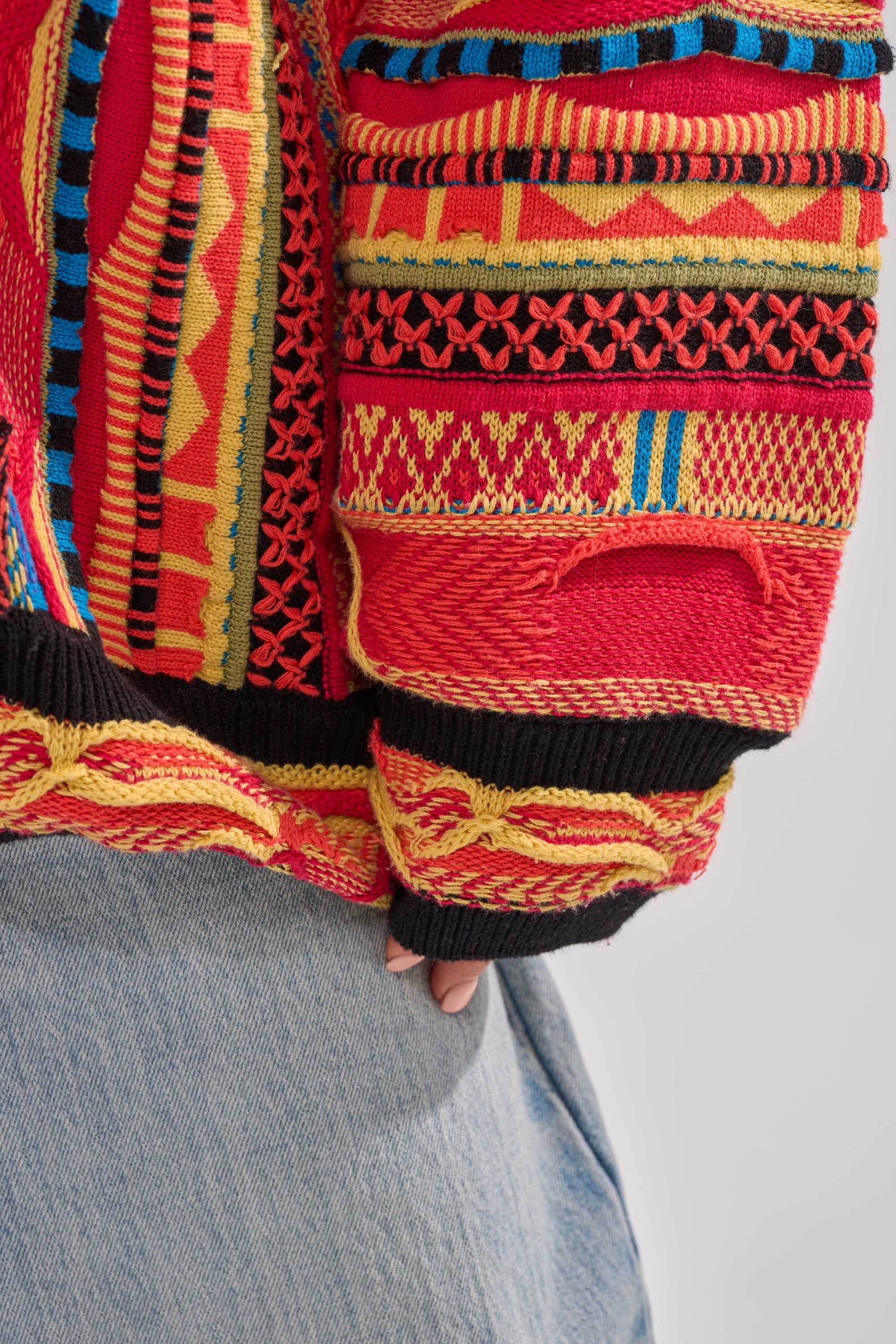 A person models the CRAZY IN LOVE CARDIGAN, a colorful retro knit sweater featuring red, yellow, and black designs, styled with light blue jeans. Only the torso and part of one hand are visible. 8