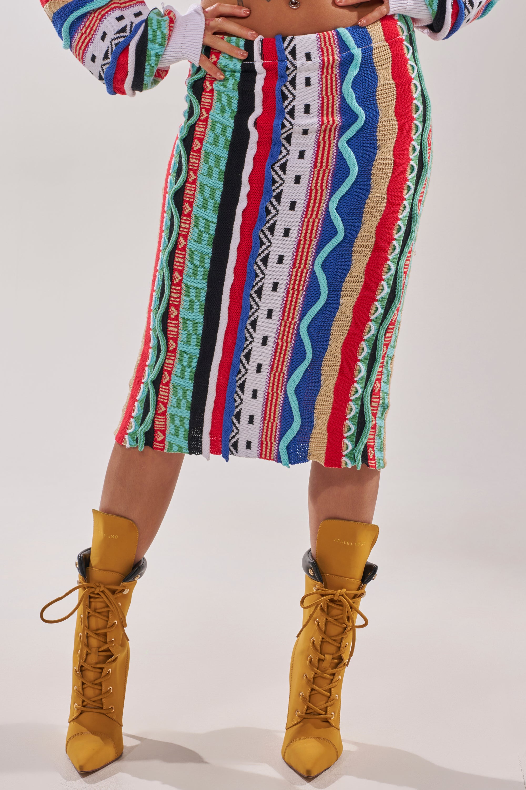 A person wearing the FEELING THE VIBES MAXI SKIRT, a multicolor knit maxi with vertical stripes, and yellow lace-up heeled boots stands against a plain light background. 1