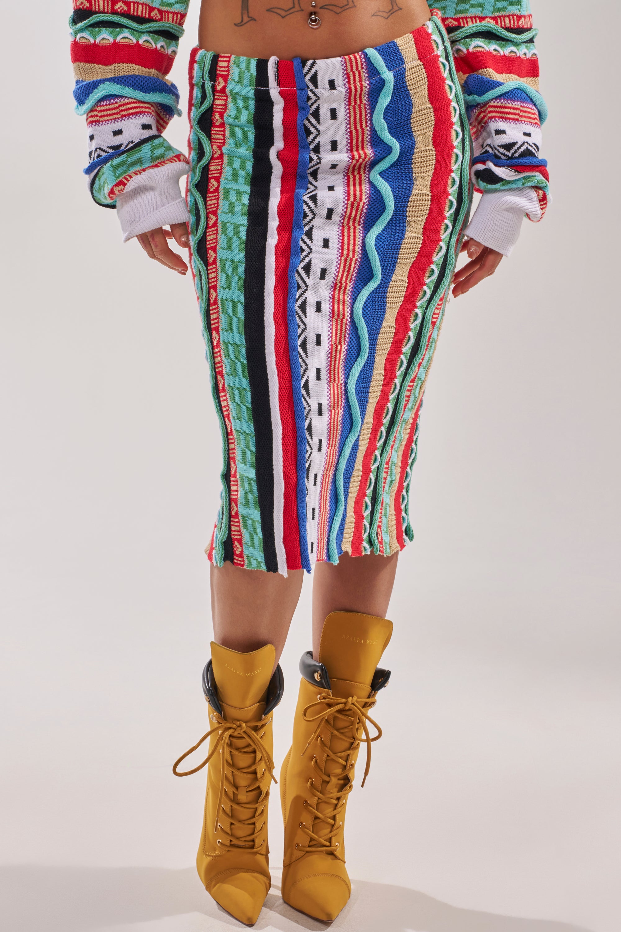 The midsection and legs of a person are shown wearing the FEELING THE VIBES MAXI SKIRT, featuring multicolor vertical stripes and patterns, paired with mustard yellow lace-up heeled boots. 2