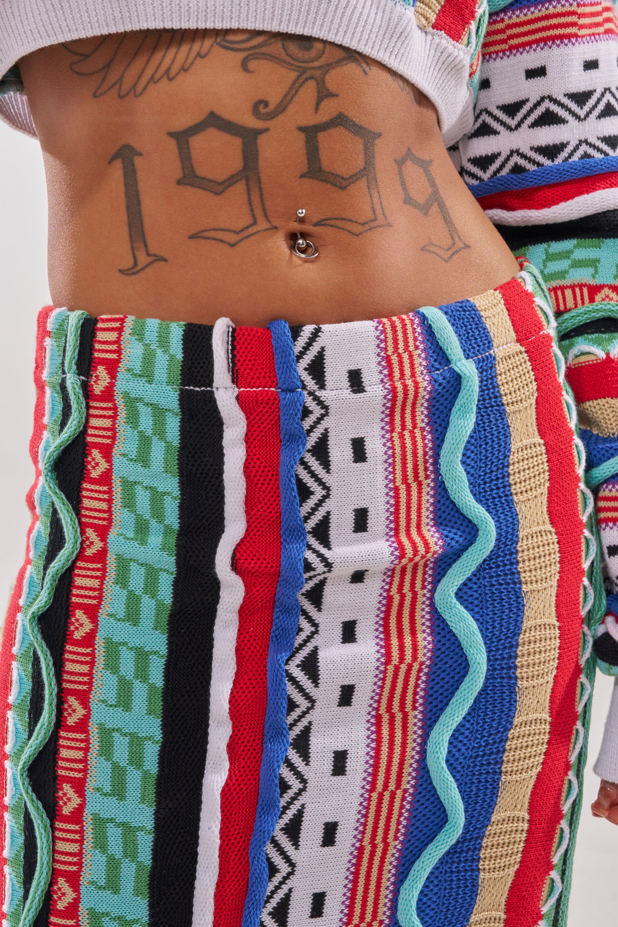 A close-up of someone wearing the FEELING THE VIBES MAXI SKIRT, featuring bold stripes and wavy patterns, showing a "1999" tattoo and belly button piercing on their exposed midriff. 8