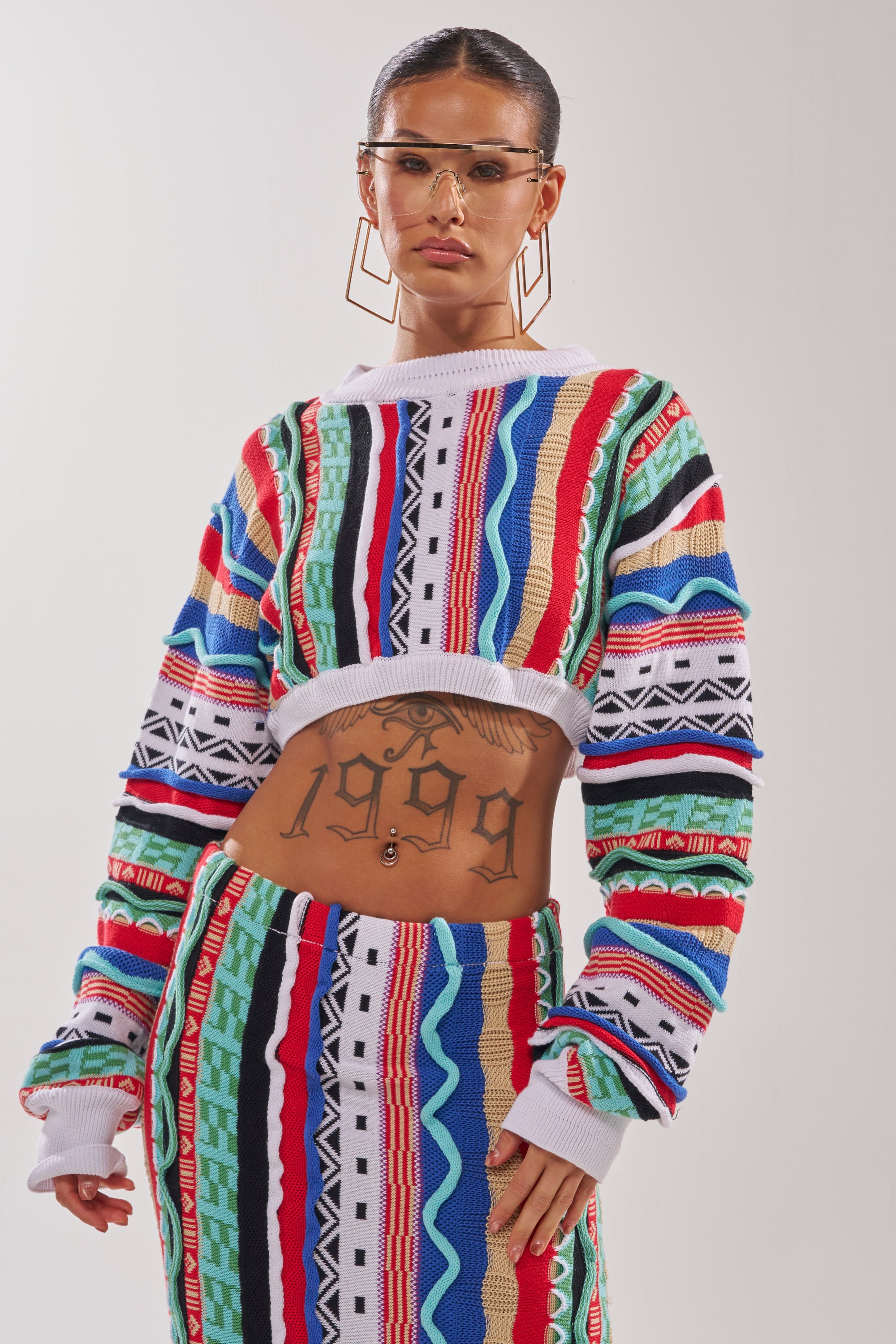 A woman poses confidently in the FEELING THE VIBES CROP SWEATER and matching skirt, accessorized with geometric glasses, hoop earrings, and a “1999” stomach tattoo, set against a plain white background. 0