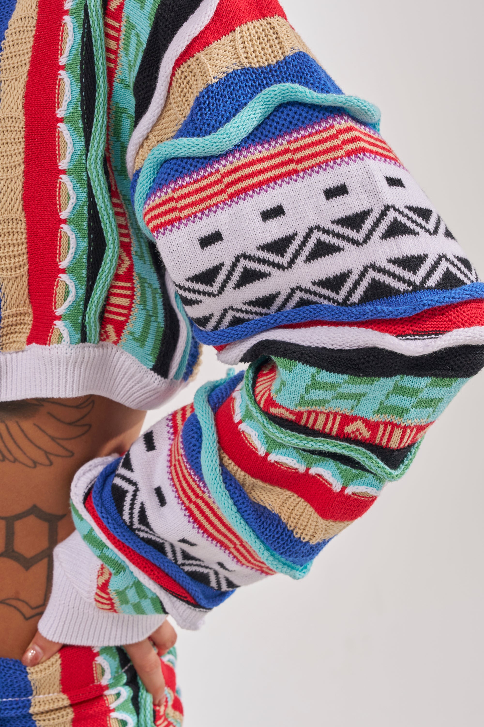 A close-up of a person wearing the FEELING THE VIBES CROP SWEATER, featuring bold multicolor geometric stripes and patterns, with its cropped fit revealing part of their tattooed torso. 9
