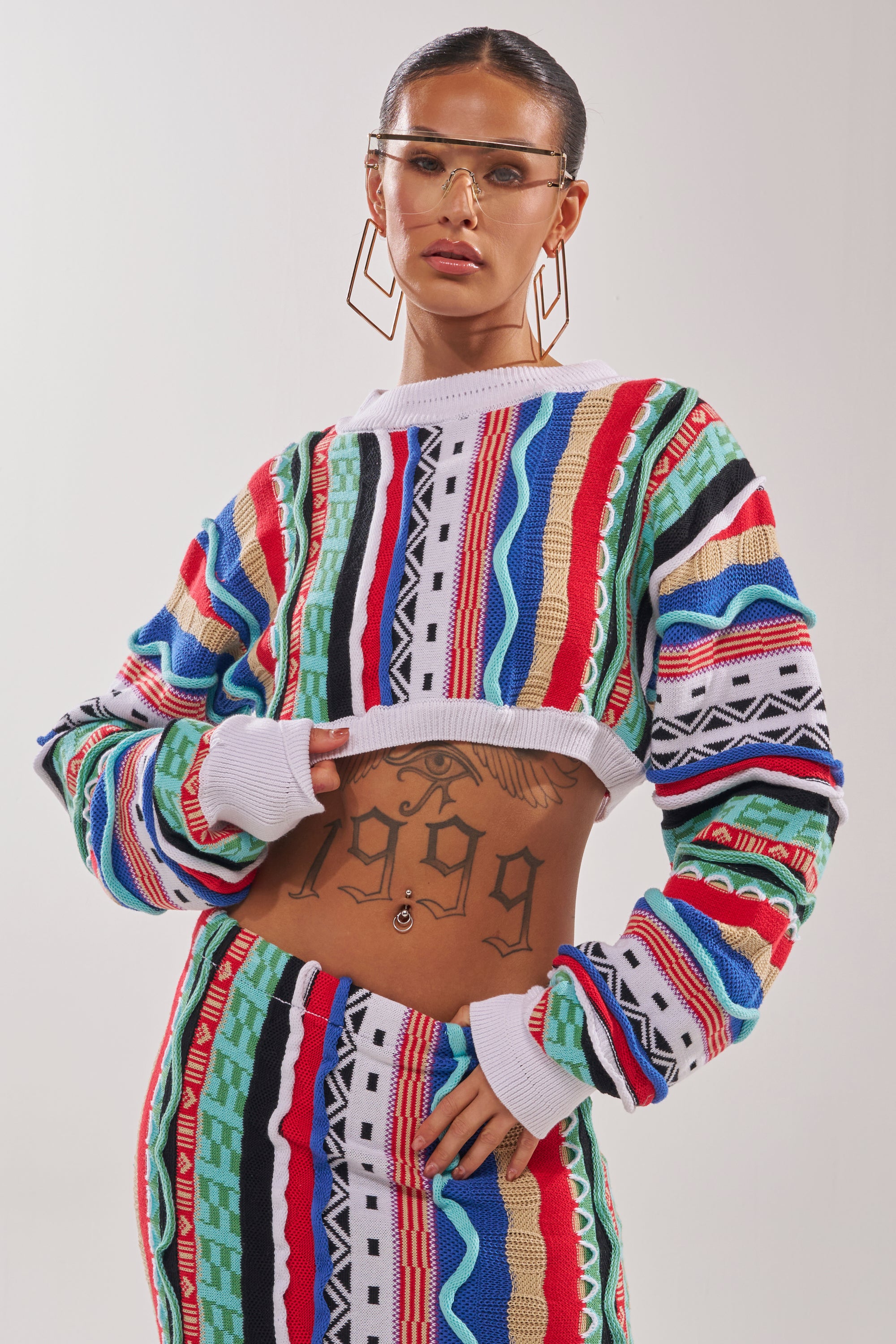 A person poses confidently against a plain background wearing clear sunglasses, bold geometric earrings, and the FEELING THE VIBES CROP SWEATER—a multicolor knit with geometric striping—revealing a "1999" tattoo on their midriff. 1