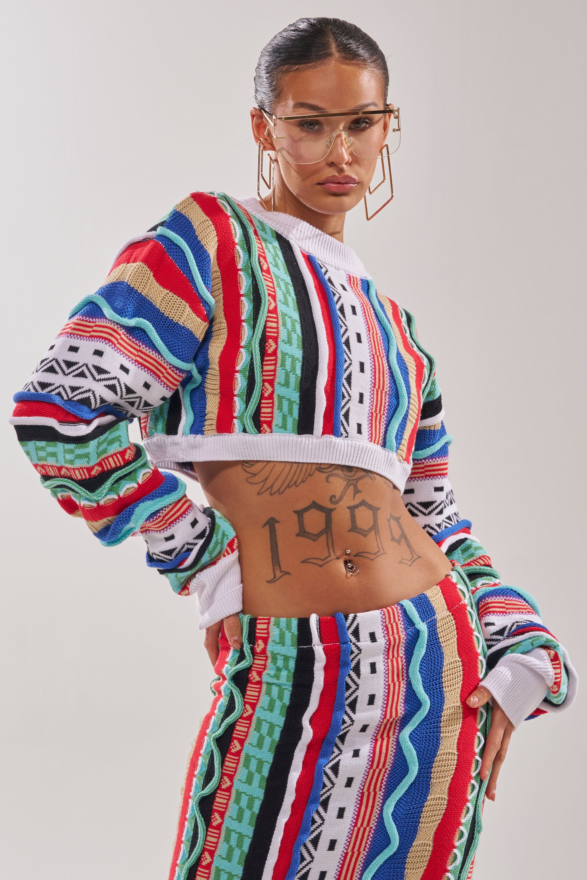 A woman with slicked-back hair and geometric glasses models the FEELING THE VIBES CROP SWEATER, a multicolor knit in a cropped fit with bold geometric stripes, paired with a matching skirt. A "1999" tattoo is visible on her stomach against a plain background. 2