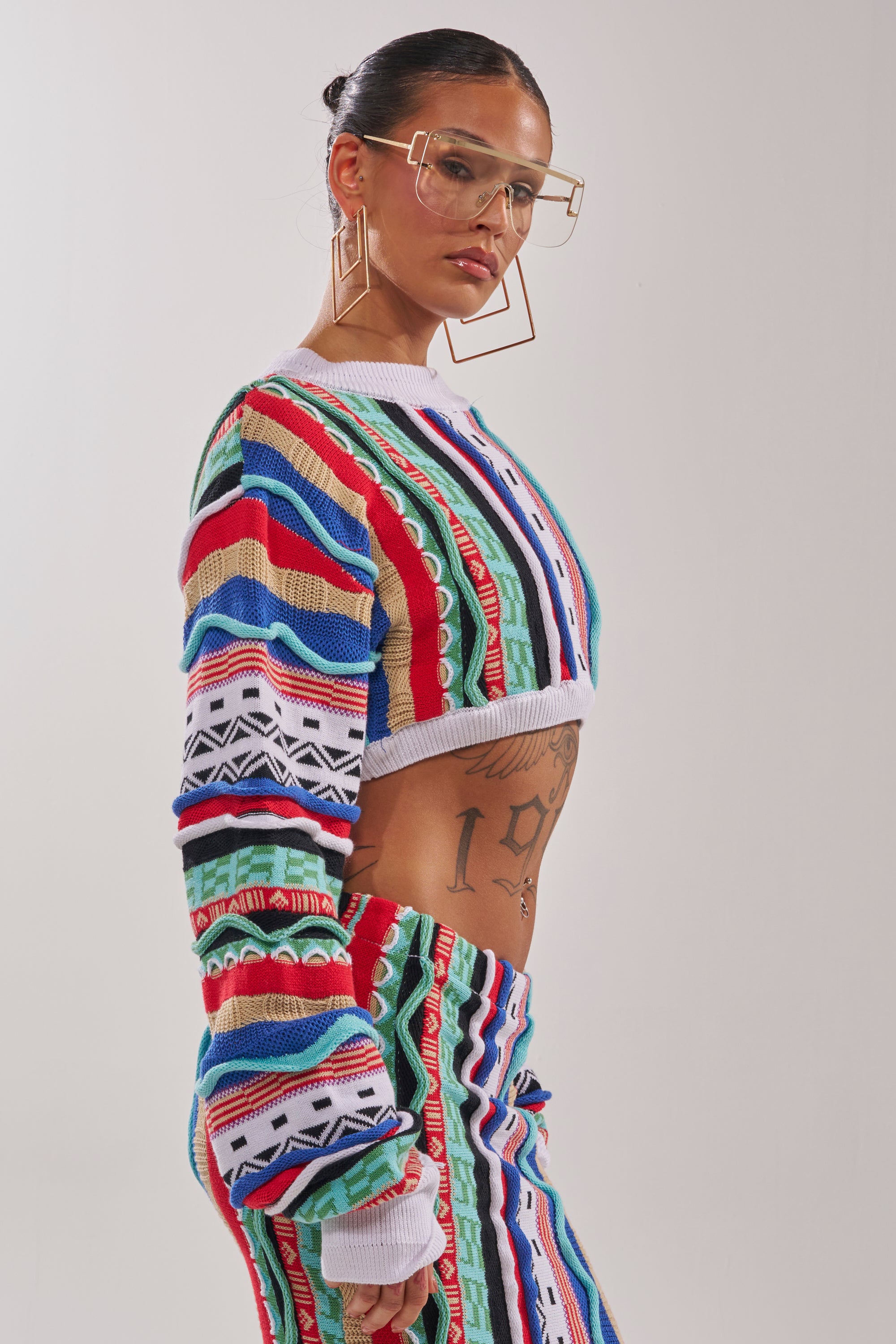 A person with slicked-back hair and bold geometric glasses poses in the FEELING THE VIBES CROP SWEATER and matching striped pants, revealing torso tattoos against a plain background. 3