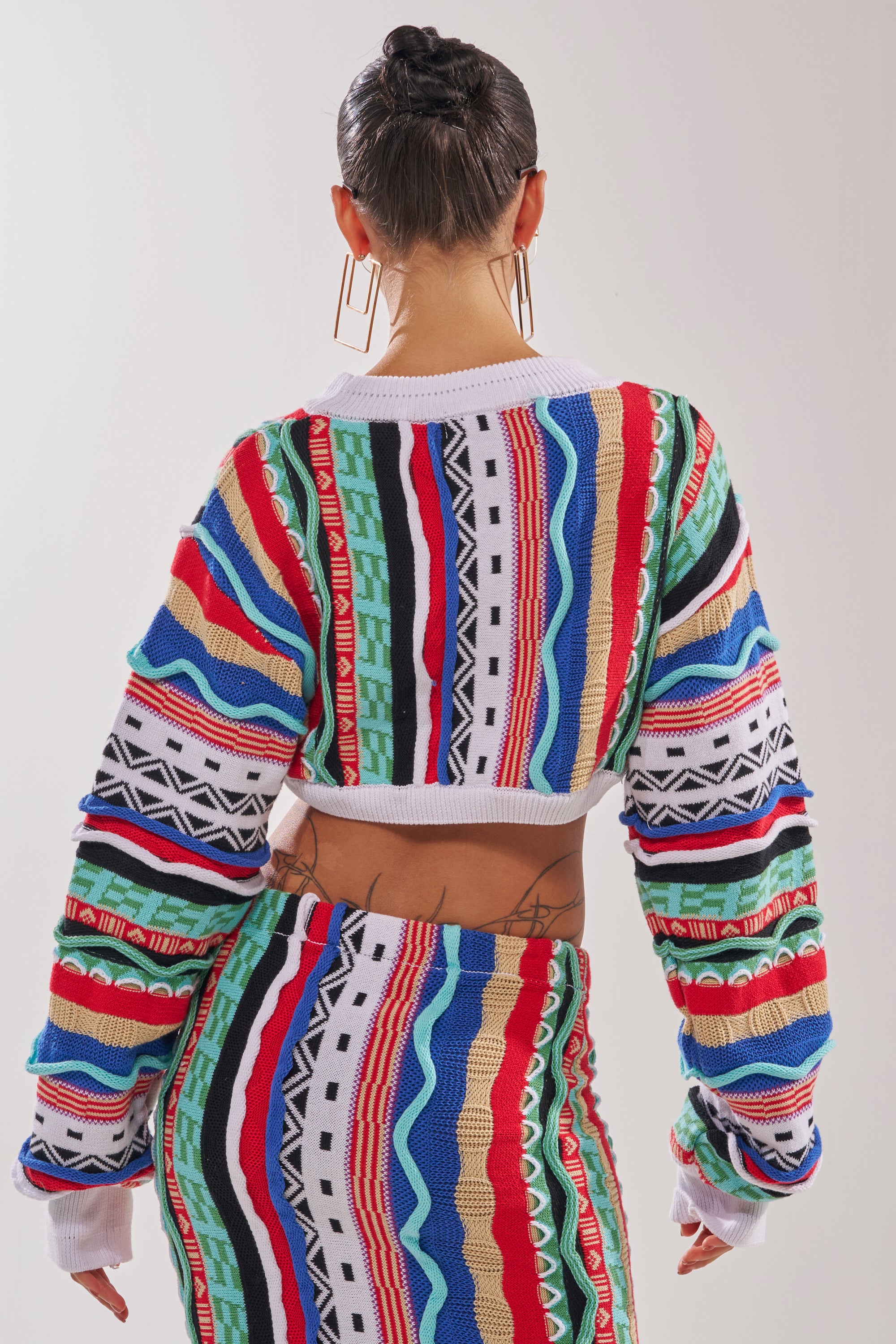 A person with slicked-back hair is seen from behind, wearing the FEELING THE VIBES CROP SWEATER with geometric stripes and vibrant patterns, paired with a matching skirt. Large hoop earrings and back tattoos complete the look. 4