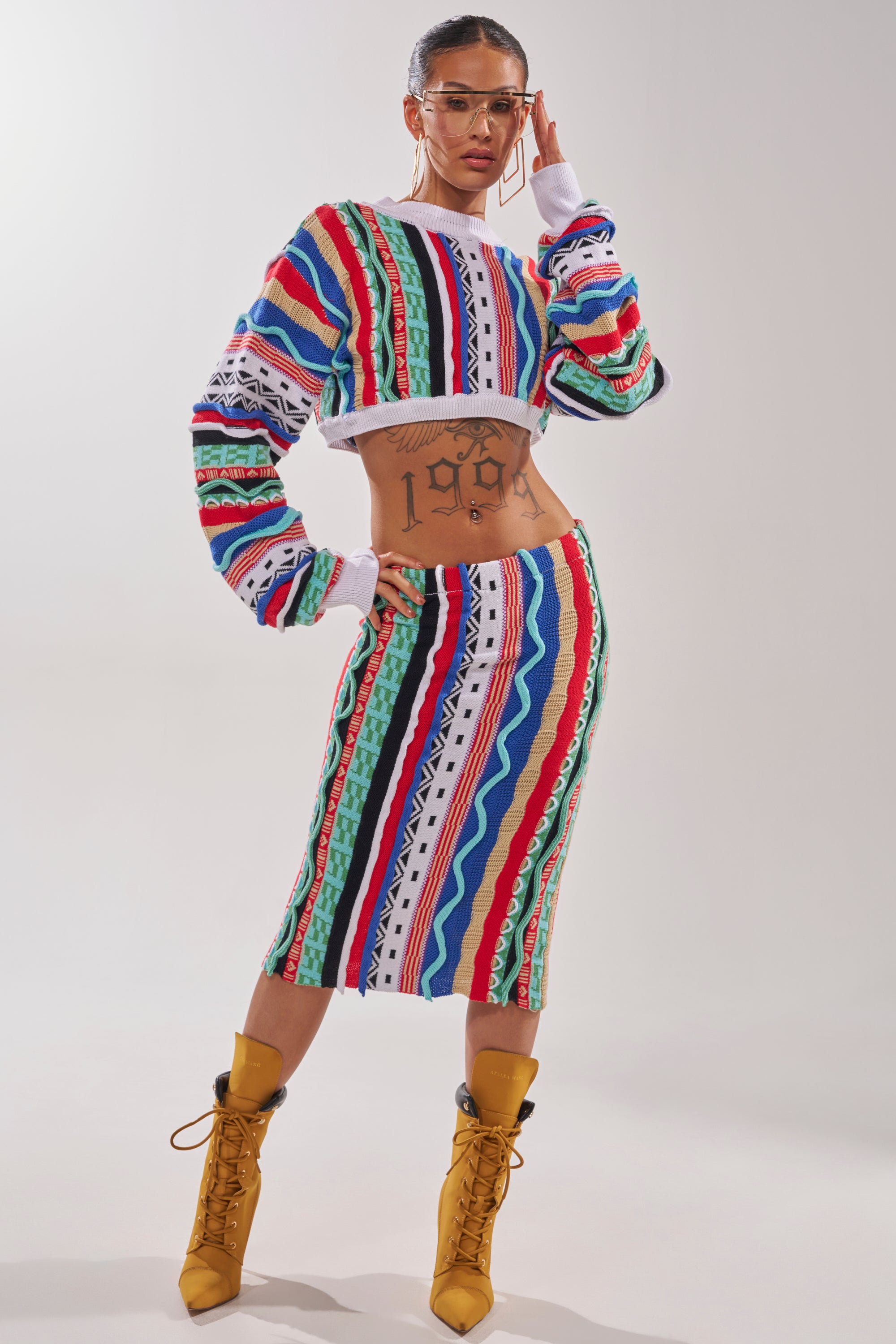 A woman in glasses and yellow heeled boots poses confidently in the FEELING THE VIBES CROP SWEATER, a multicolor geometric knit, paired with a matching pencil skirt. Her midriff is exposed, revealing a large "1997" tattoo on her stomach. 5