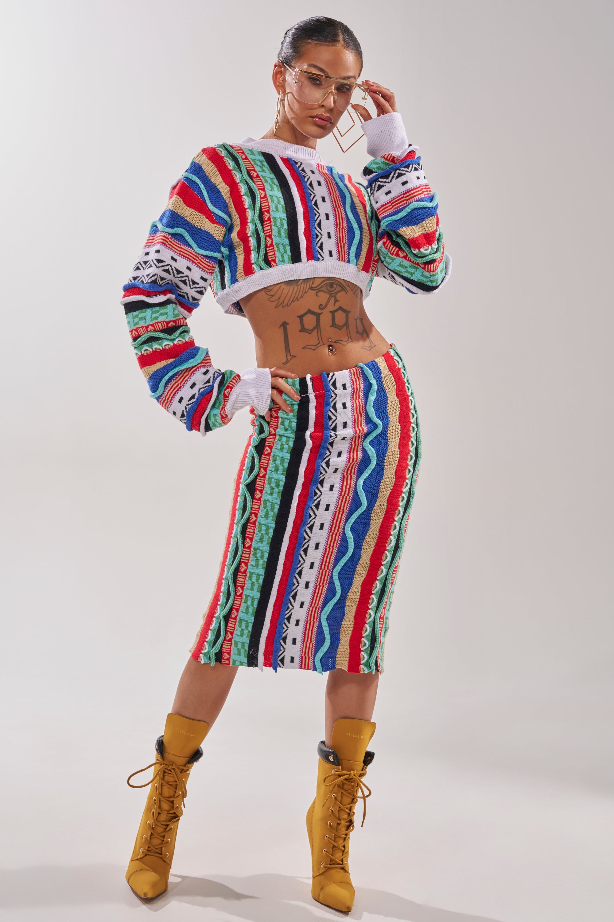 A woman poses confidently in the FEELING THE VIBES CROP SWEATER, a multicolor geometric knit, paired with a matching high-waisted skirt. She completes the look with clear glasses, large hoop earrings, and yellow lace-up ankle boots. 6