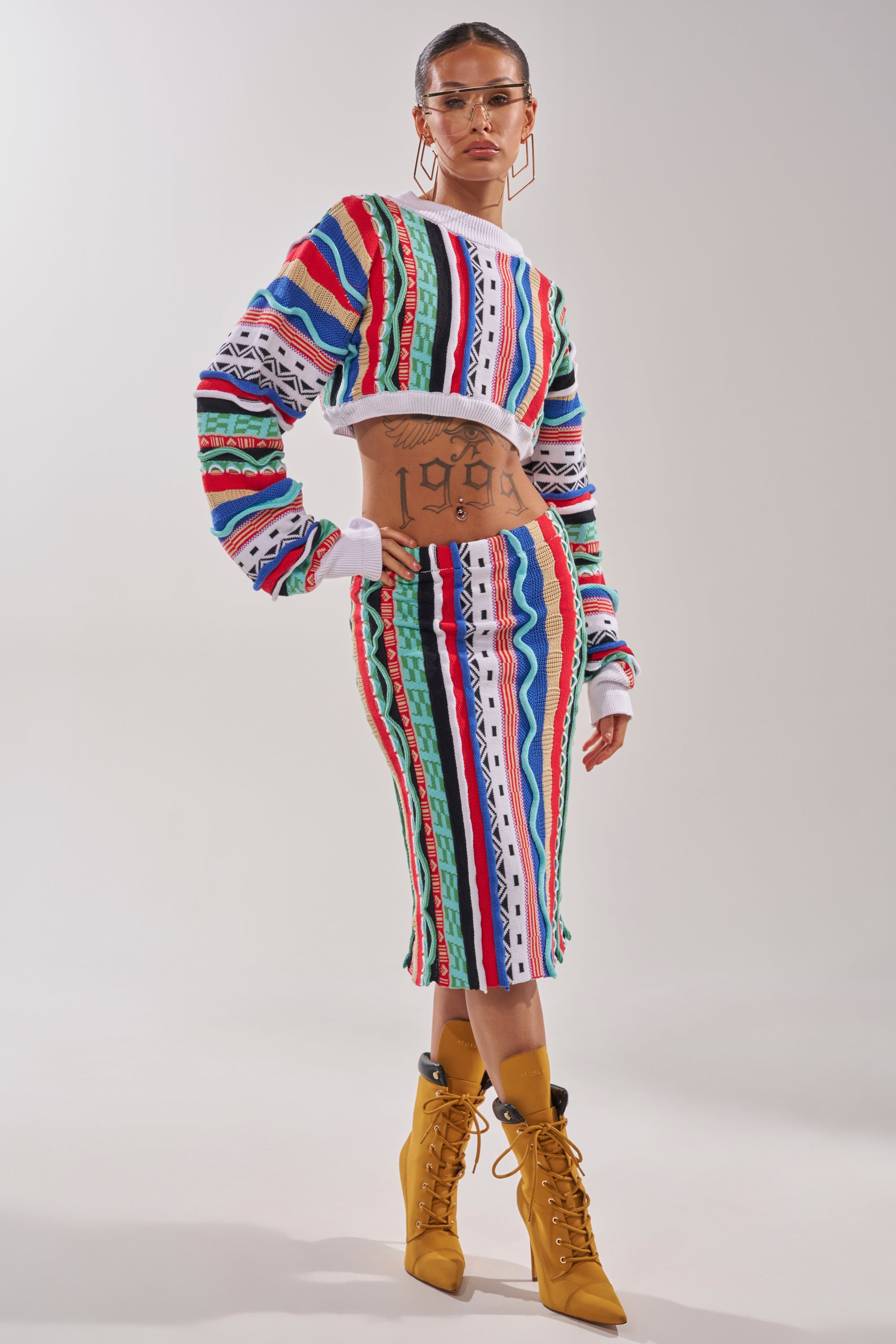 A woman in the FEELING THE VIBES CROP SWEATER and a matching multicolor geometric striped skirt, with tattoos, clear glasses, and gold lace-up heeled boots, stands facing forward against a plain light background. 7