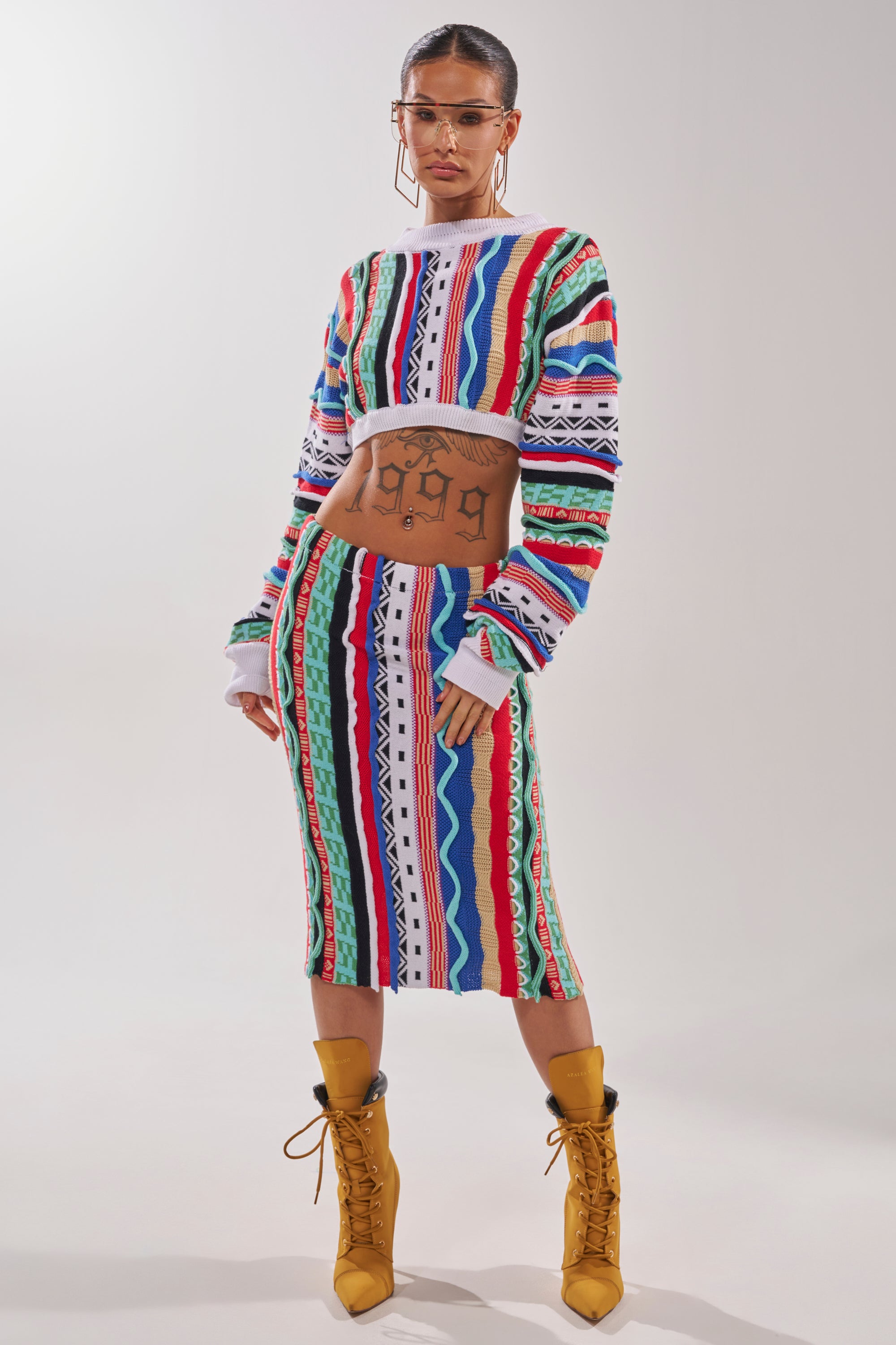 A person in the FEELING THE VIBES CROP SWEATER with geometric striping, a matching skirt, yellow lace-up boots, hoop earrings, and glasses stands against a plain background, showing a "1999" tattoo on their midriff. 8