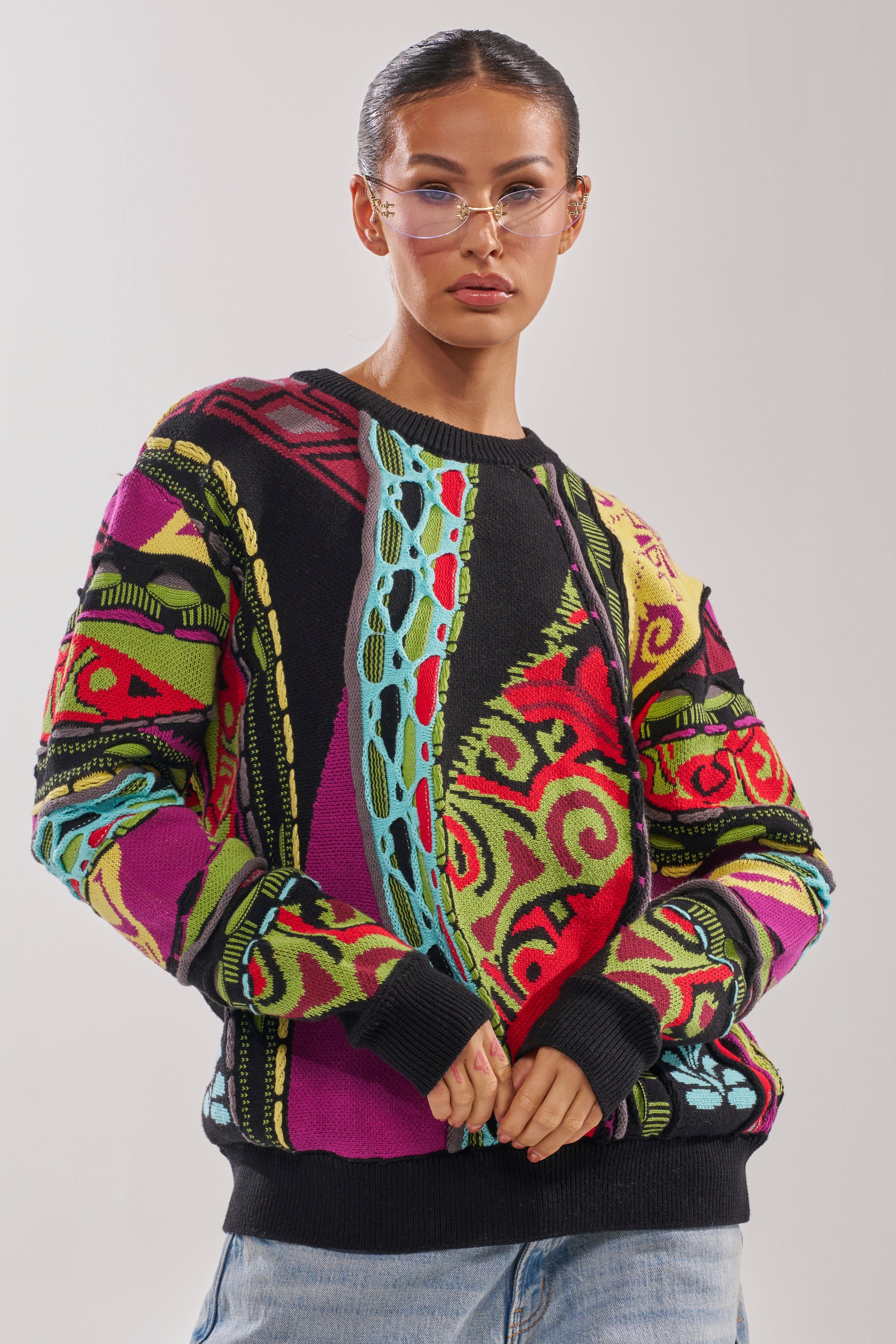 A person wearing the GET GROOVY SWEATER—an oversized vintage piece with red, green, blue, and purple designs—and clear glasses stands confidently against a plain light background. 0