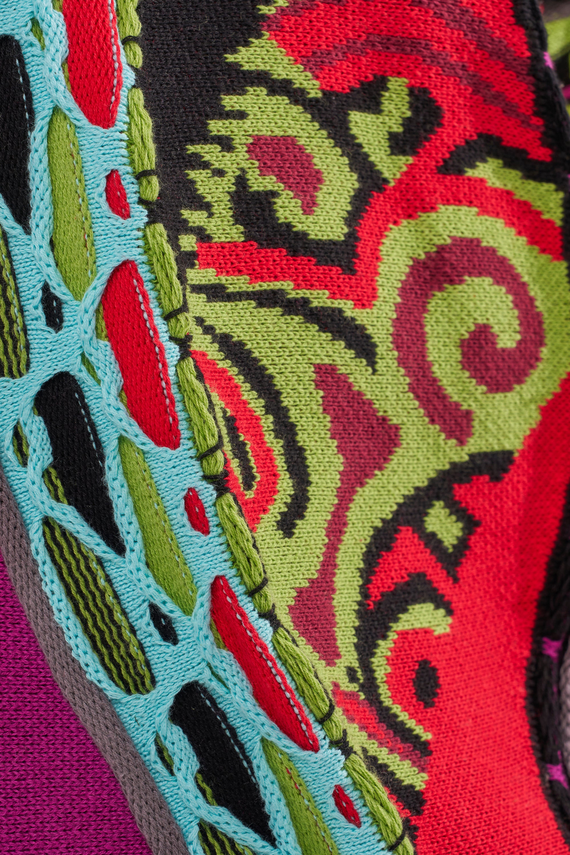 Close-up of the GET GROOVY SWEATER, featuring vibrant red, green, and black swirls and a decorative edge with teal and black embroidery in an open, lattice-style knit. 8
