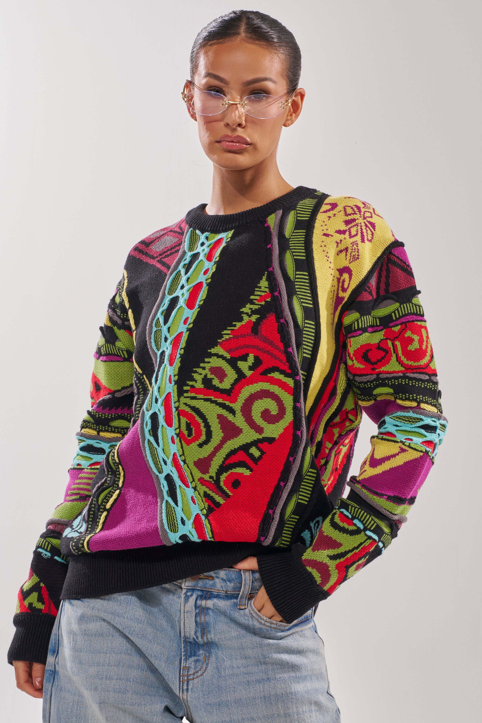 A person in clear glasses, light blue jeans, and the GET GROOVY SWEATER—an oversized vintage knit with bold multicolor graphics—stands against a plain light gray background. 1