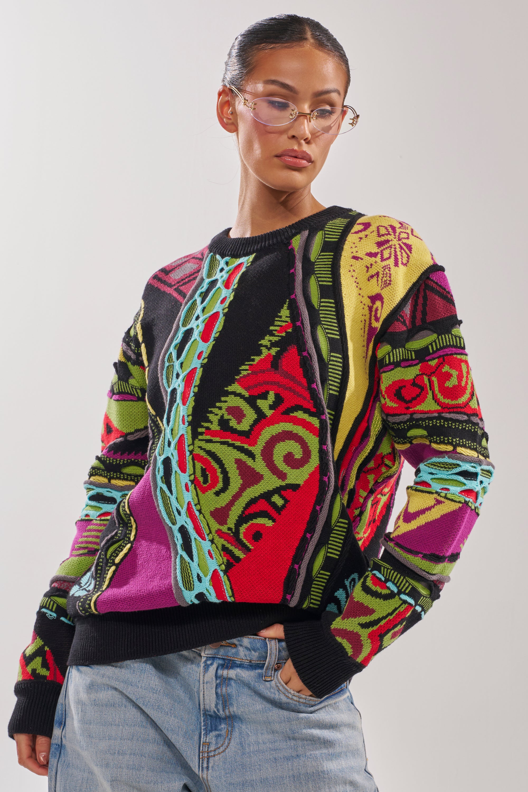 A person wearing glasses and the GET GROOVY SWEATER, a vintage-inspired knit with colorful geometric and abstract designs, paired with light blue jeans, stands against a plain light background. 2
