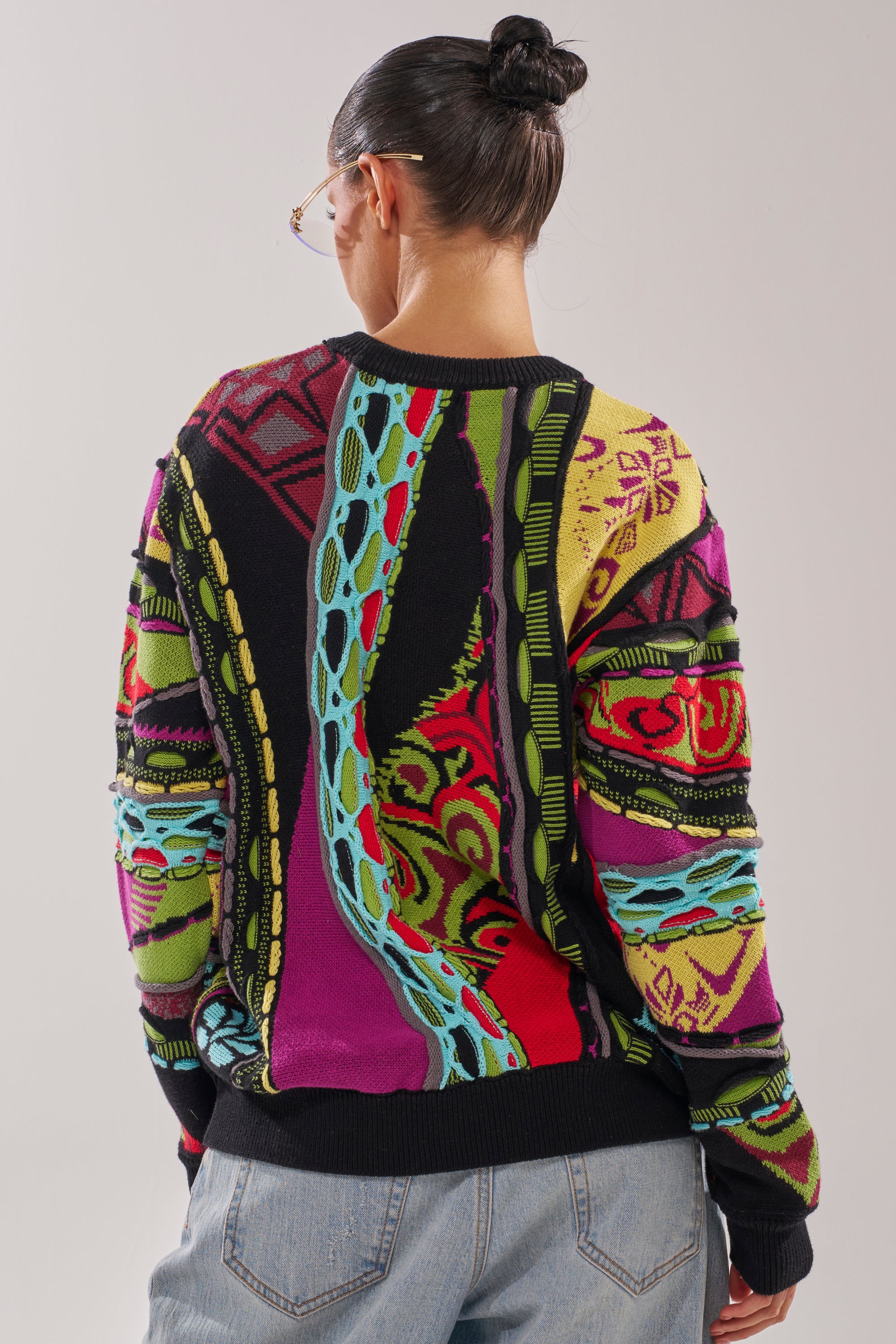 A person with dark hair in a bun, wearing sunglasses, blue jeans, and the colorful GET GROOVY SWEATER with abstract designs in red, yellow, green, blue, and purple is shown from the back against a plain background. 4