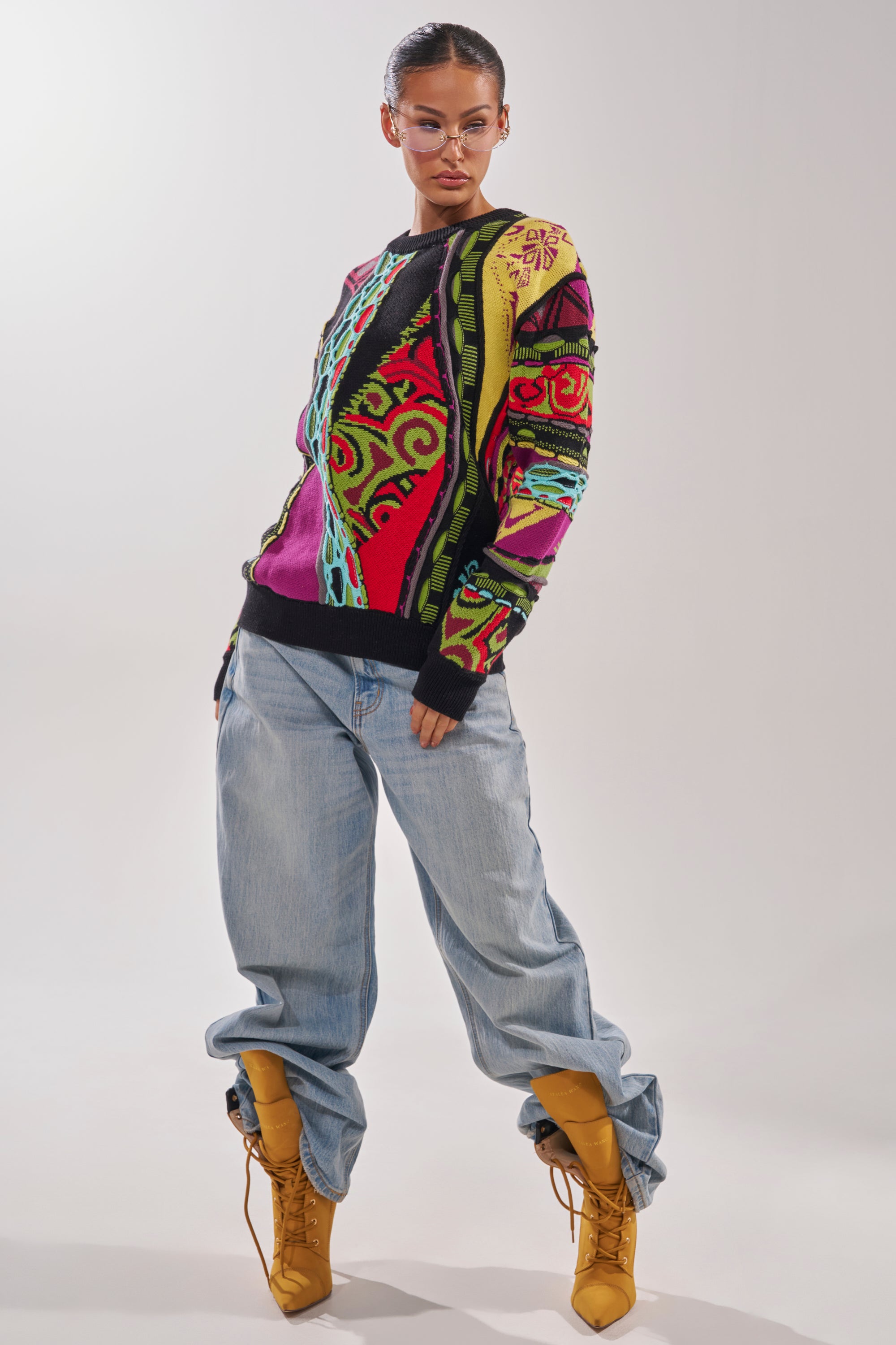 A person poses against a plain light background wearing the GET GROOVY SWEATER, vintage-inspired with graphic knits, paired with loose light blue jeans, yellow lace-up boots, and clear glasses. 6