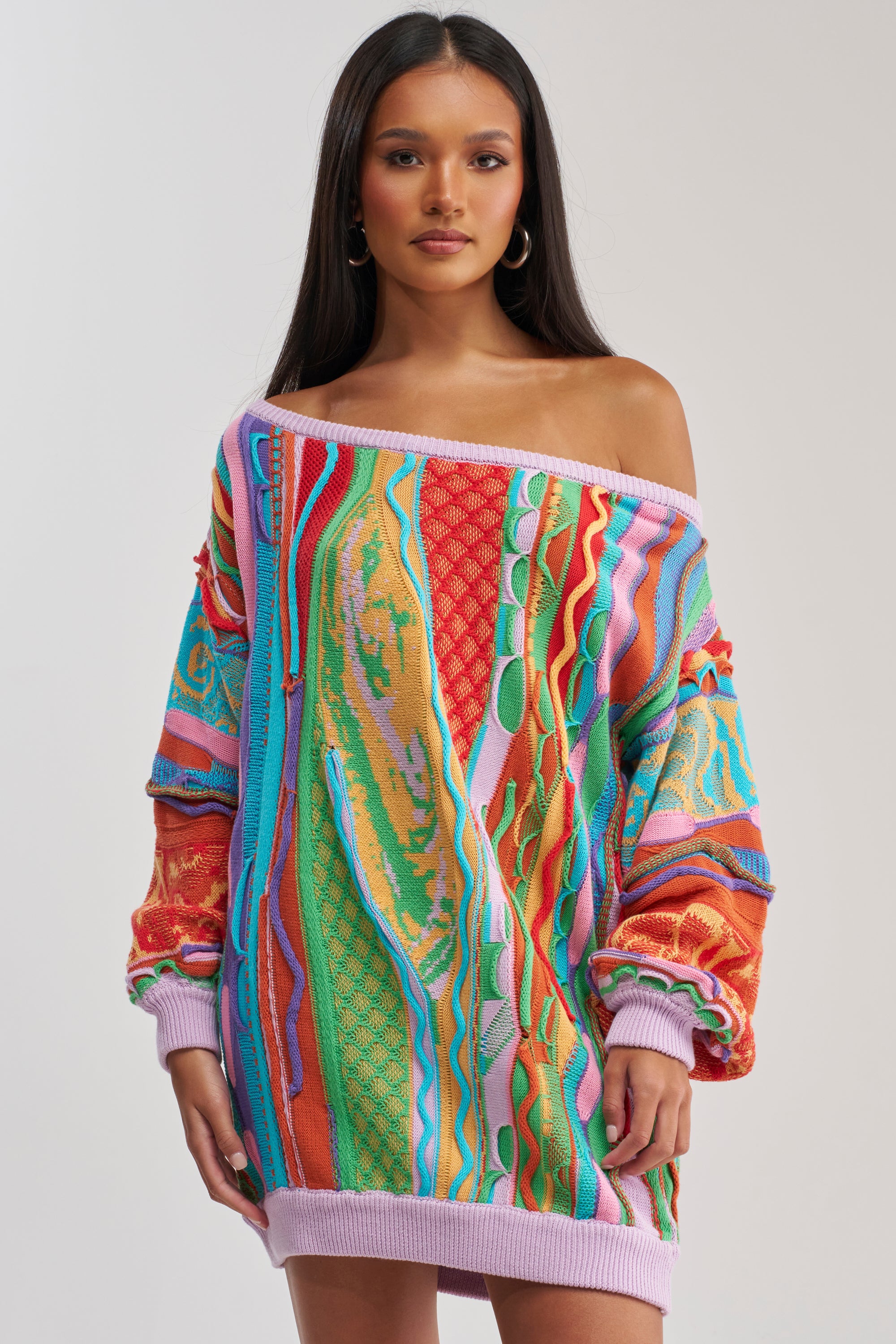 A woman models the CROWD FAVORITE SWEATER MINI DRESS—an oversized, off-the-shoulder piece with bold, textured red, orange, green, blue, and purple patterns—standing against a plain light background for vibrant winter style.