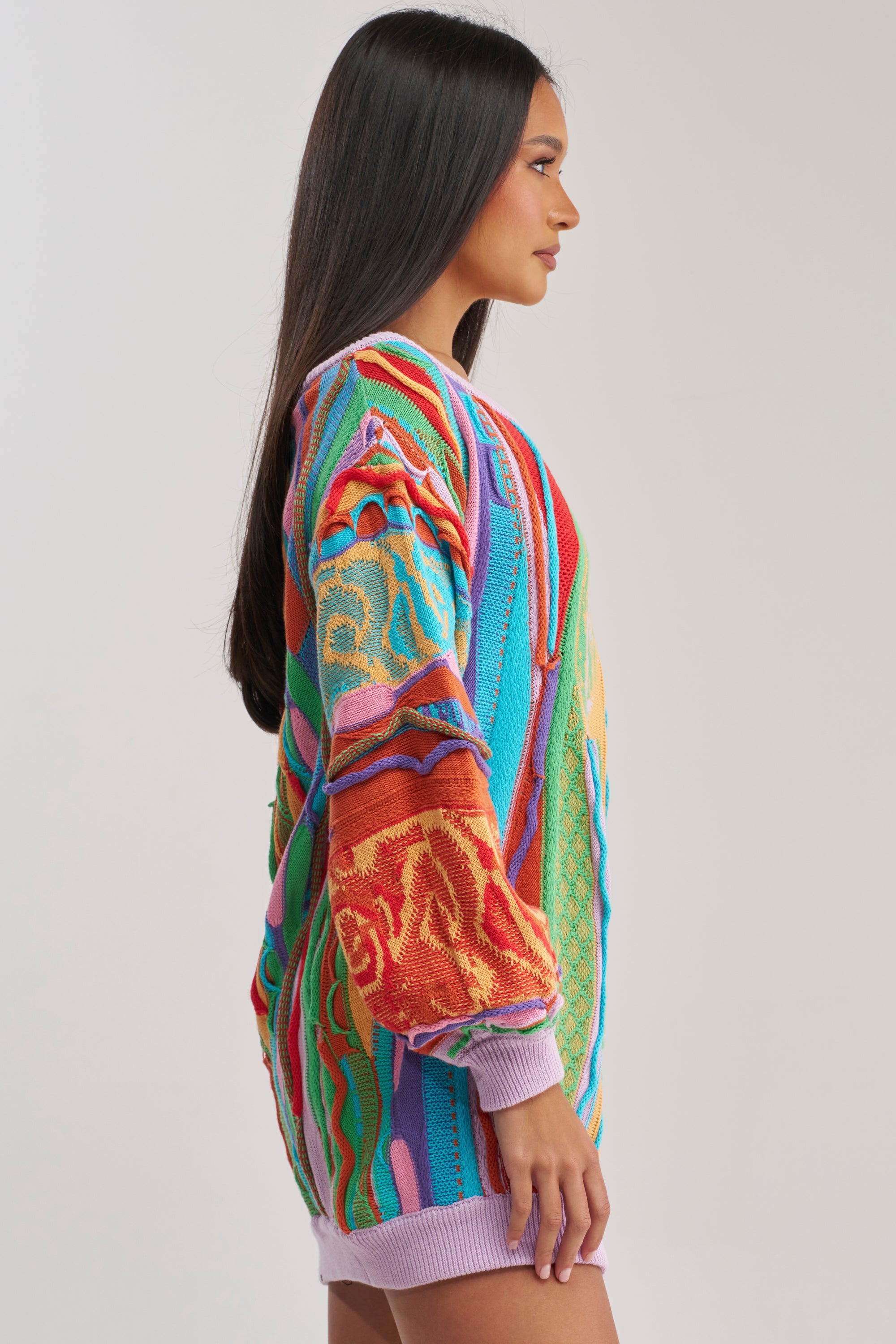 A woman with long dark hair stands in profile, wearing the CROWD FAVORITE SWEATER MINI DRESS—a vibrant knit dress with abstract patterns and lilac cuffs—perfect for winter layering, against a plain light gray background.