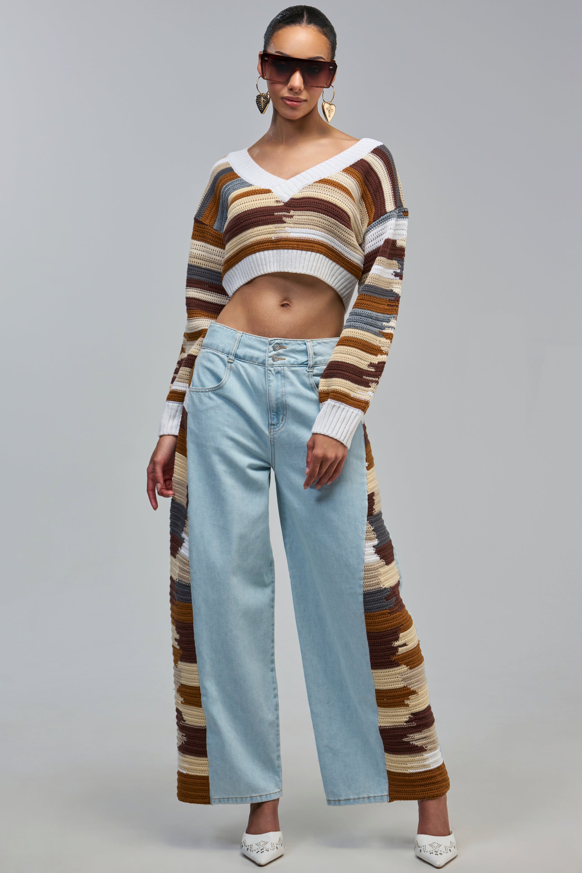 A person wears a cropped, striped knit sweater and DRIVING ME INSANE DENIM WITH CROCHET PANEL jeans, featuring light-wash denim with brown, cream, and white crochet accents.