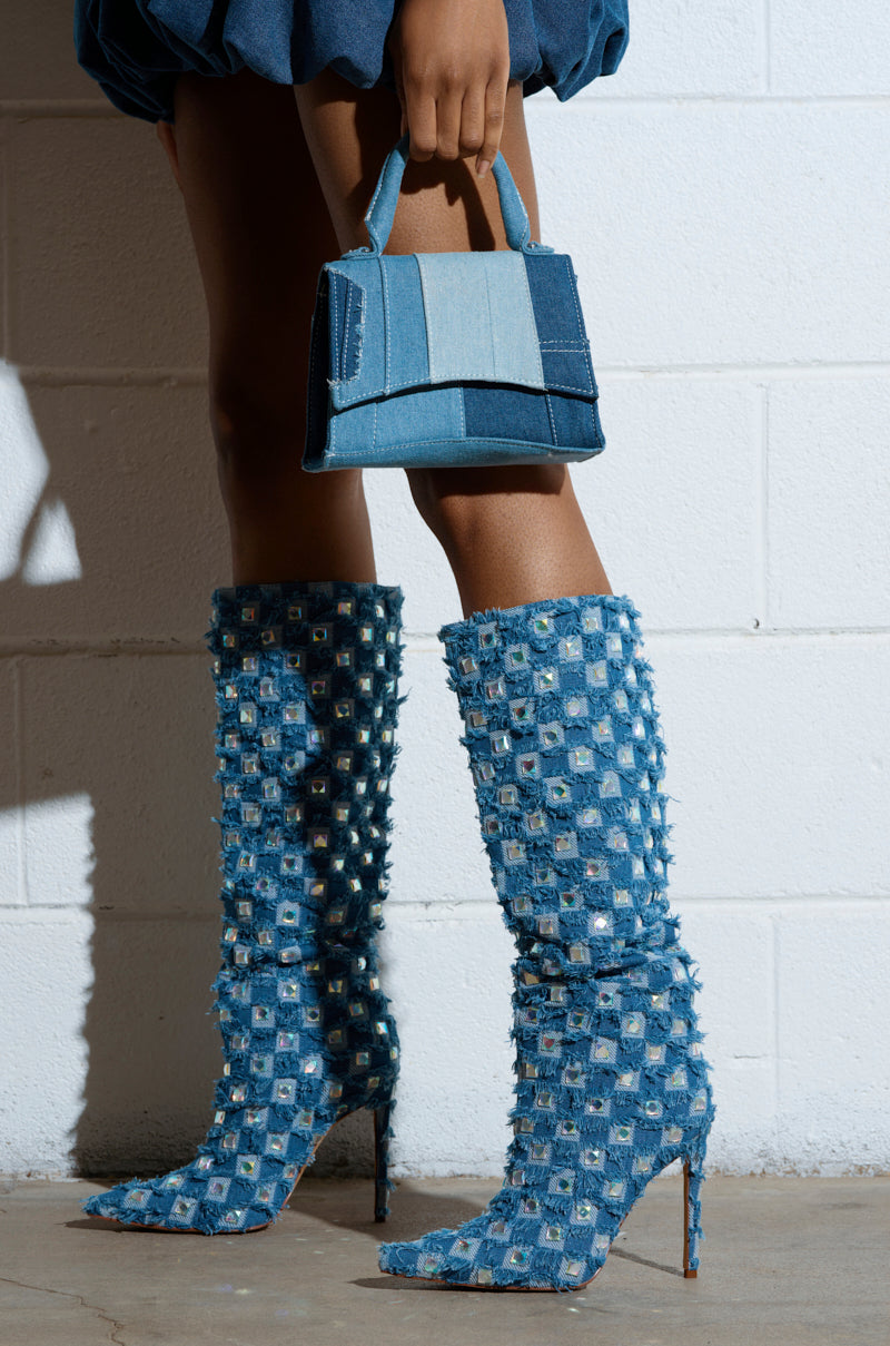 A person wearing AZALEA WANG EVE denim knee-high boots with rhinestone-embellished stilettos and a matching embellished small handbag stands against a white brick wall. 1