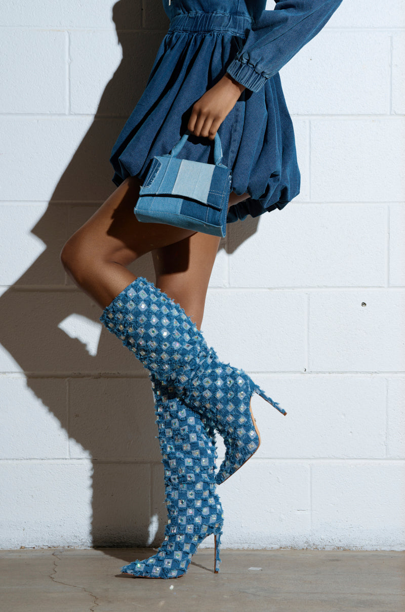 A person in a denim dress holds a denim patchwork handbag. Their AZALEA WANG EVE Denim Knee High Embellished Boots, with stiletto heels, add flair to their outfit as rhinestone details glisten against a white brick wall. 2