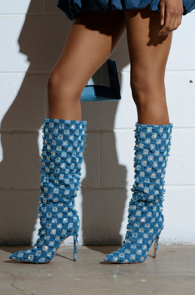 Against a white brick wall, a stylish figure stands on a concrete floor wearing AZALEA WANG EVE denim knee-high embellished boots with square patterns and stiletto heels. They elegantly clutch a blue purse as shadows play on the backdrop. 0