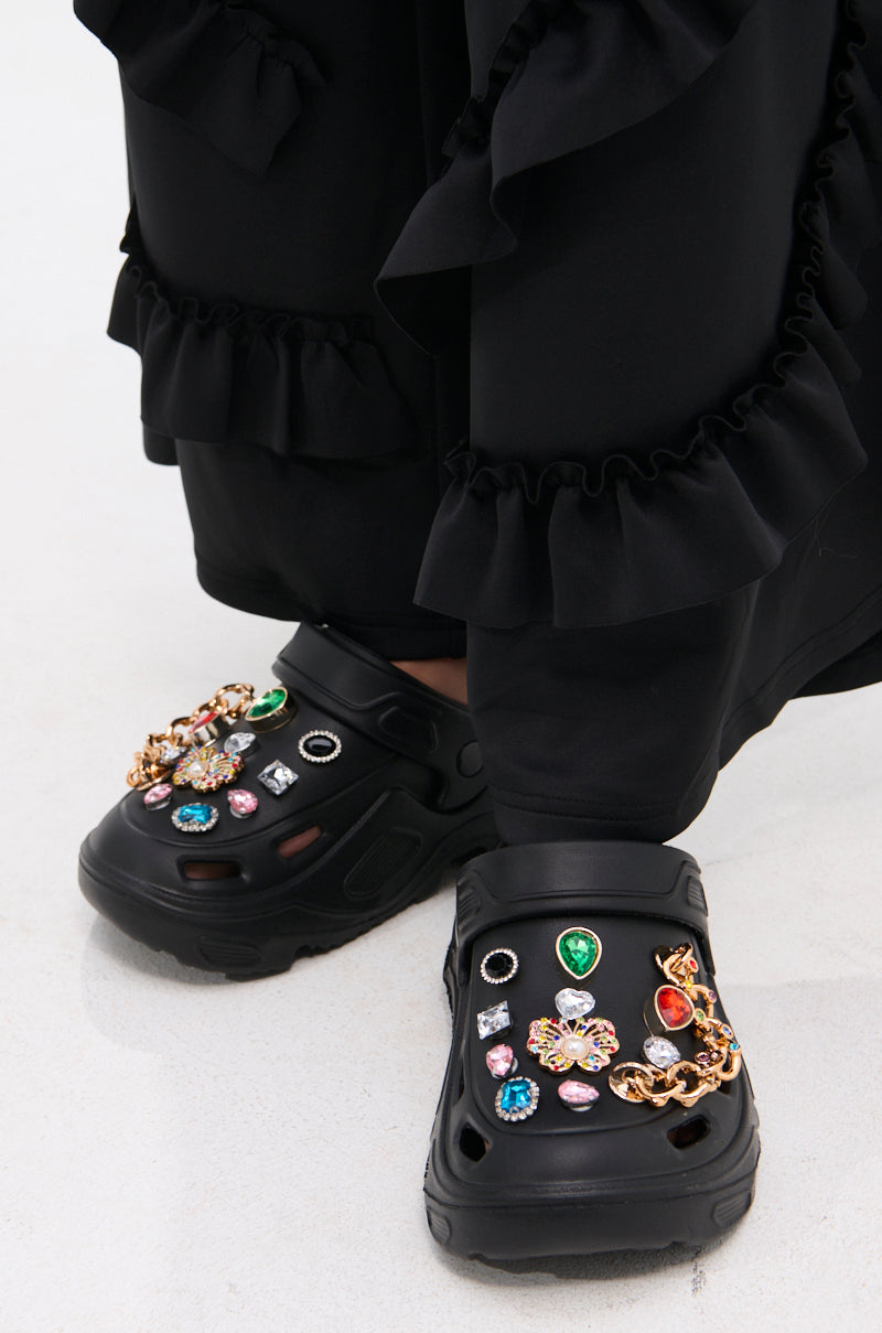 Close-up of the AZALEA WANG EVERETT BLACK MULTI STONE GOLD CHAIN FLAT, styled with black ruffled pants, featuring black multi-stone rhinestone charms and gold chains on a white floor.