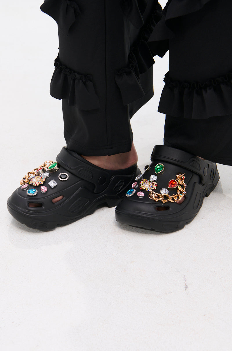 A person wearing black ruffled pants and AZALEA WANG EVERETT BLACK MULTI STONE GOLD CHAIN FLAT shoes with colorful jeweled charms, standing on a light-colored floor.
