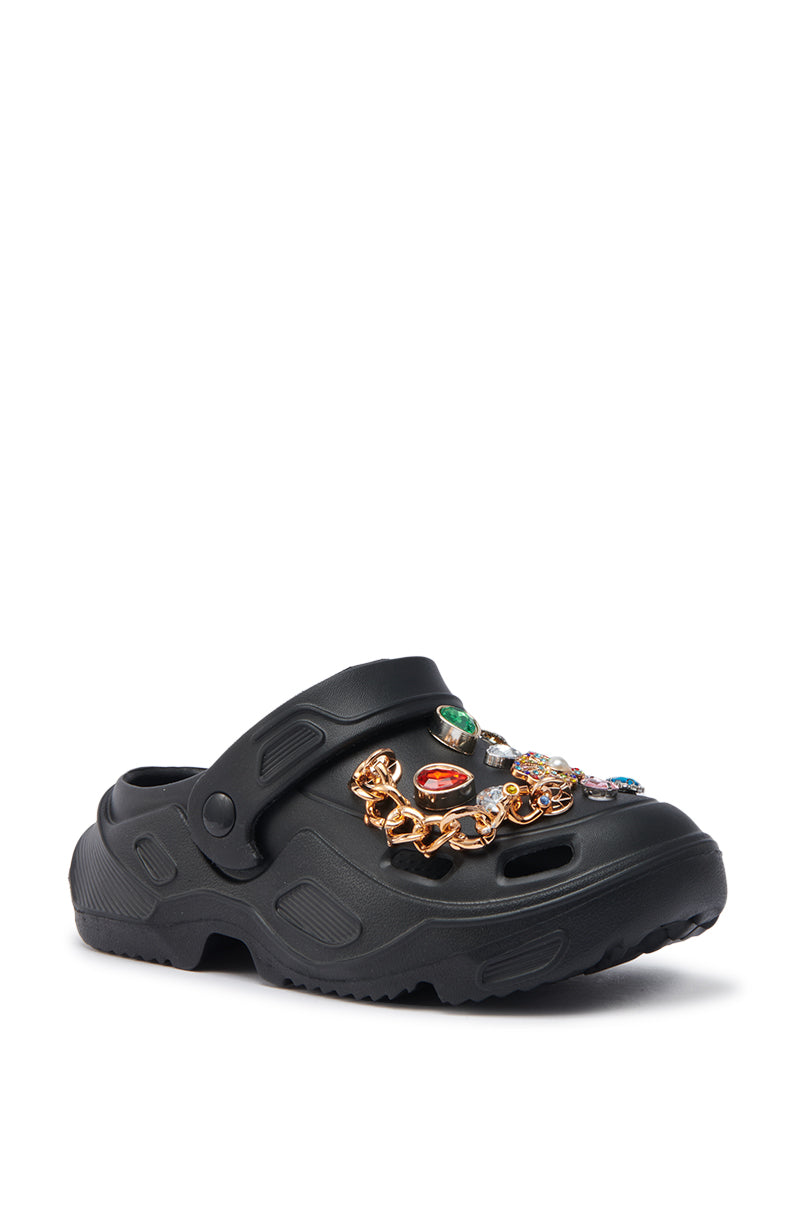 The AZALEA WANG EVERETT BLACK MULTI STONE GOLD CHAIN FLAT is a black slip-on clog with a back strap, chunky sole, and embellished upper featuring colorful stones and gold chain details for a standout look.