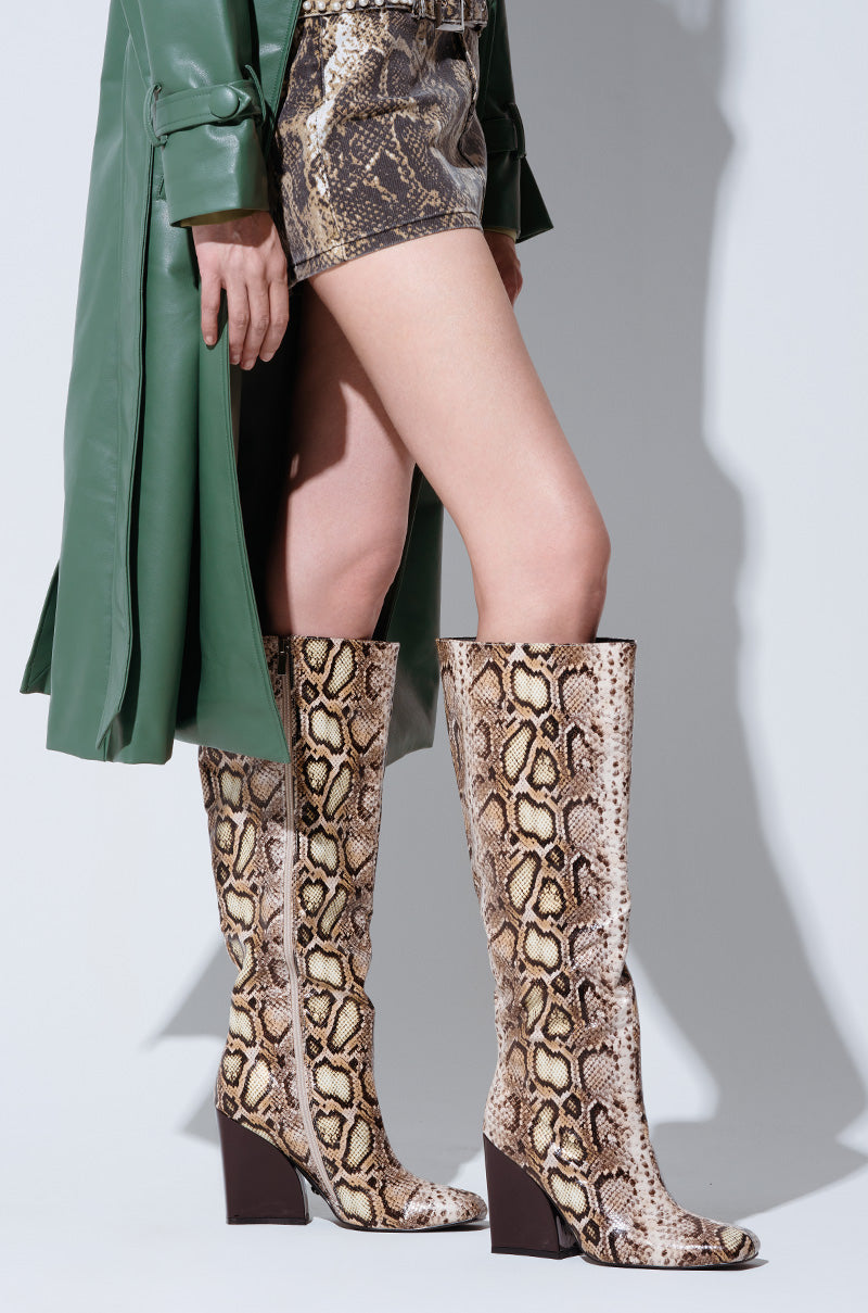 A person wearing a green trench coat and a snakeskin-patterned mini skirt, paired with the AZALEA WANG EVERGREEN MULTI SNAKE BOOT featuring knee-high faux snakeskin with a chunky wedge heel. The shadow is visible against a light background.