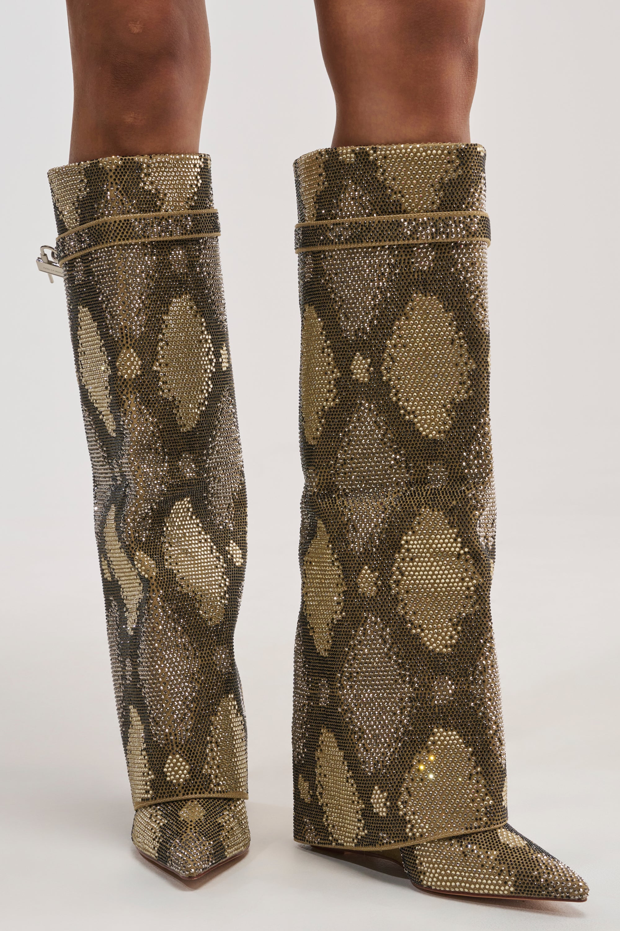 A close-up view of a person wearing the AZALEA WANG EVO KHAKI SNAKE SPARKLE WEDGE BOOT, a knee-high style with pointed toes and dazzling rhinestone embellishments arranged in a brown and gold snakeskin pattern, is shown against a white background. The distinctive boot features a wedge heel and an eye-catching fold-over cuff design for statement-making style.
