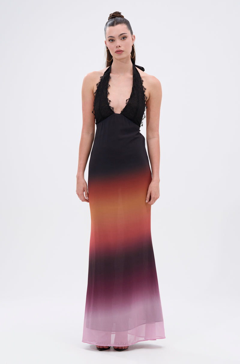 A woman faces forward in the SUNSET DREAMING OMBRE MAXI DRESS, featuring a plunging halter neckline, black lace top, and a maxi skirt that fades from black to amber to pink. Her straight dark hair is styled in a high ponytail.