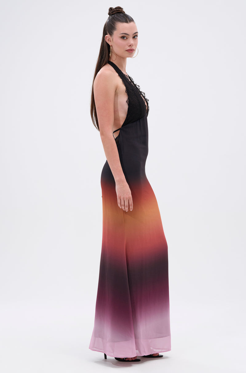 A woman stands in profile wearing the SUNSET DREAMING OMBRE MAXI DRESS, featuring a plunging halter neckline, black-to-purple ombre hues with red and orange shades, gauzy unlined fabric, and her long hair styled half-up.