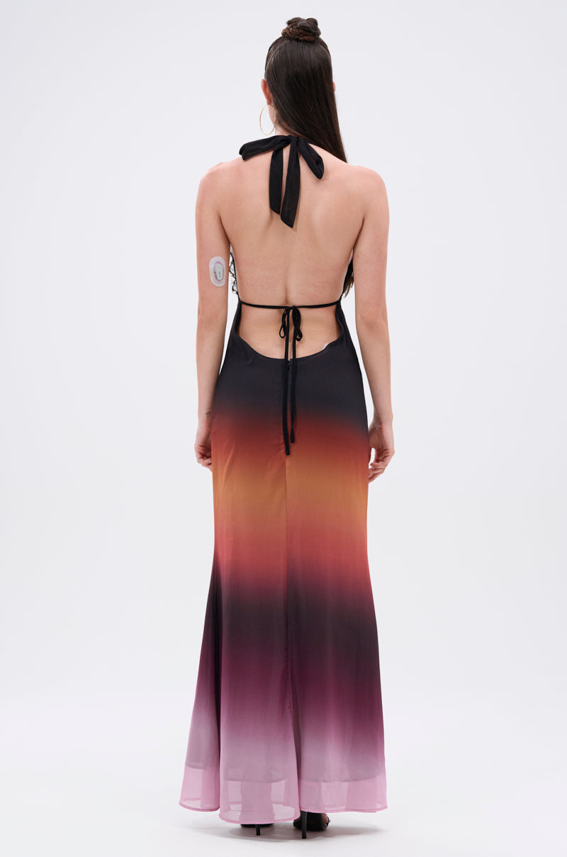 A woman with long brown hair and a visible glucose monitor on her left arm wears the SUNSET DREAMING OMBRE MAXI DRESS, featuring a plunging halter neckline and a gauzy unlined fabric that fades from black to orange and purple.