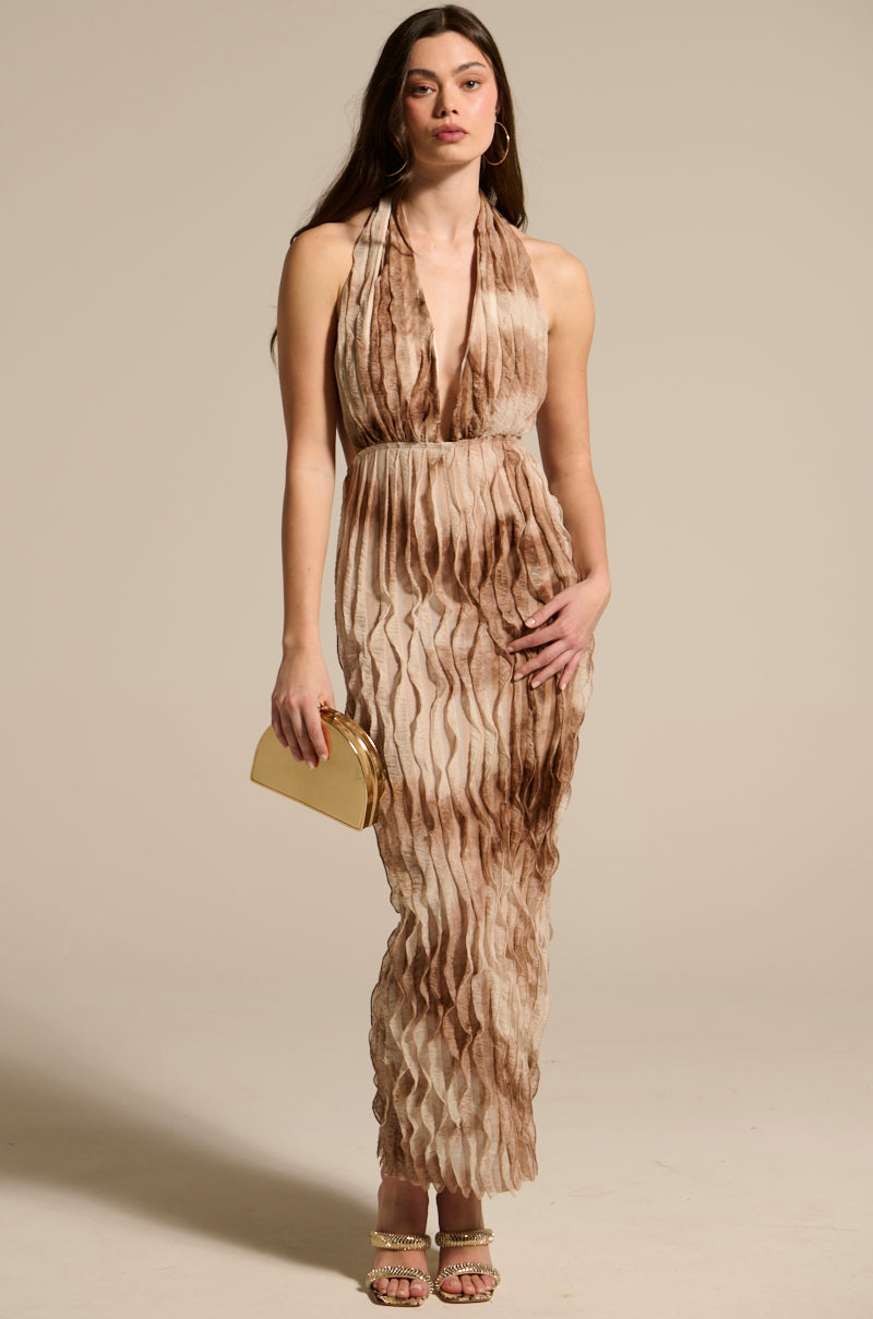 A woman wears the LOOK BUT DON'T TOUCH PRINTED MAXI DRESS—a sleeveless, floor-length piece with a beige and brown tie-dye print and deep V-neckline. She accessorizes with a gold clutch, hoop earrings, and heeled sandals against a neutral background.