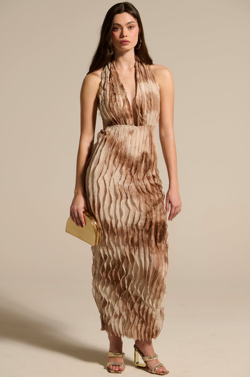 A woman wears the LOOK BUT DON'T TOUCH PRINTED MAXI DRESS, featuring a halter neckline and pleated, wavy texture in beige and brown. She accessorizes with a gold clutch, gold heels, and hoop earrings.
