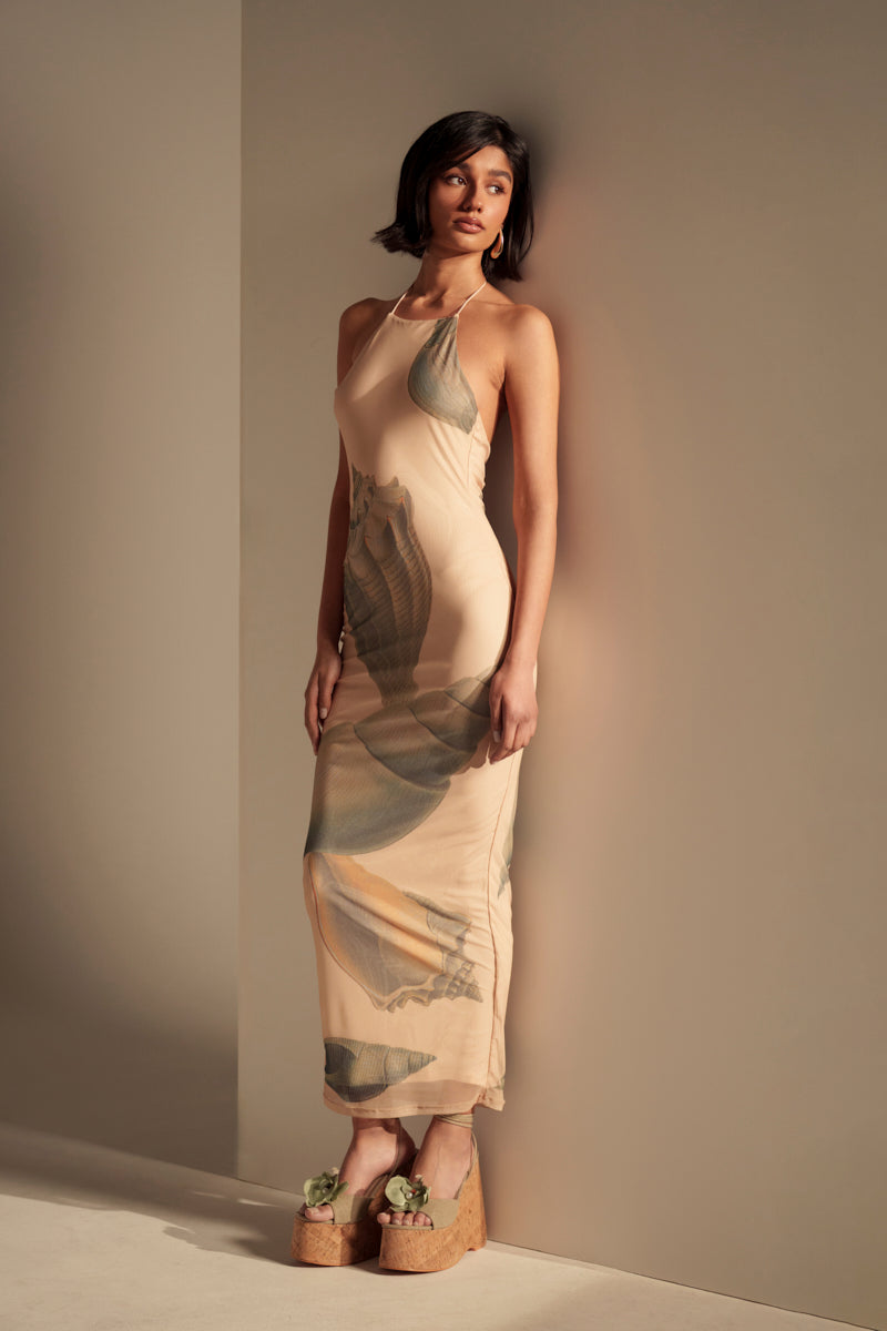 A woman with short dark hair leans against a beige wall in a minimal, softly lit space, wearing the BY THE SEASHORE PRINTED MAXI DRESS—a sleeveless, fitted maxi dress with abstract leaf patterns.