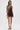 A woman poses against a white background wearing the ALL ABOUT ME LEOPARD PRINT MINI DRESS, a sleeveless, open-back mini dress with a cowl neckline. She completes the look with a metallic purse and ankle-strap high-heeled sandals.