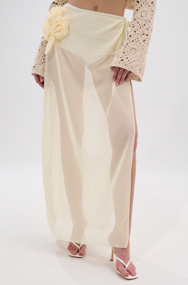 Wearing the CROCHET BEAUTY COVER UP SKIRT in sheer pale yellow with a large flower at the waist, styled with a beige crochet sleeve top and white heeled sandals, this beach-to-brunch look stands out against a plain background. 0