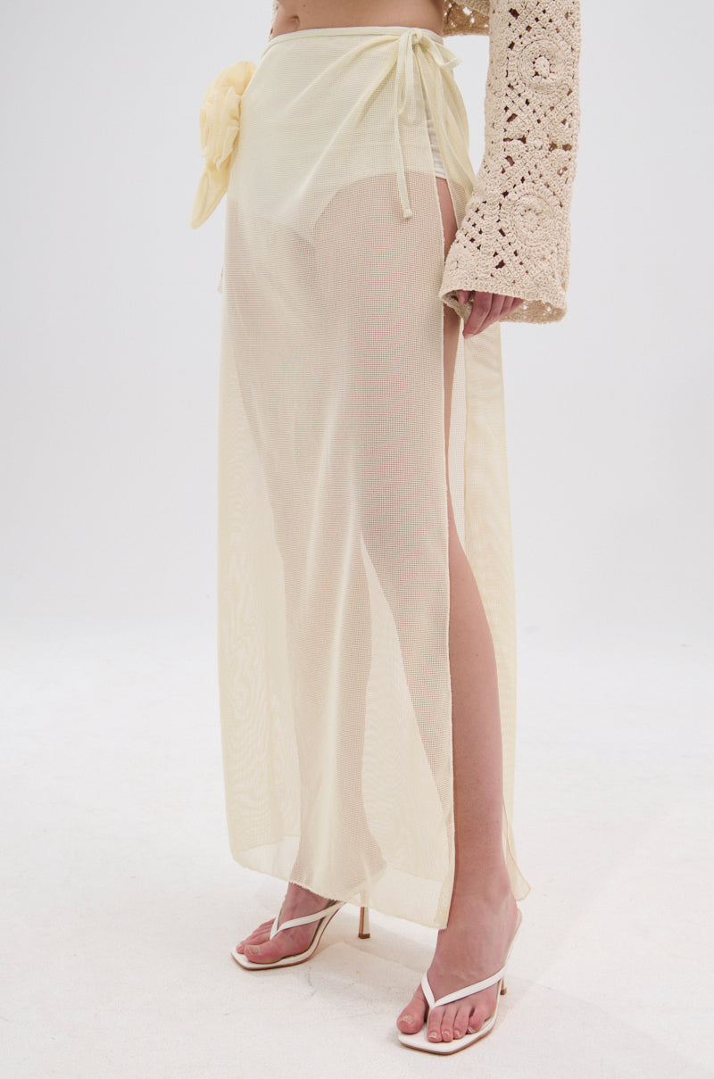 A person wears the CROCHET BEAUTY COVER UP SKIRT—a sheer, light yellow wrap skirt with a side tie and floral accents—paired with a textured beige long-sleeve top and white heeled sandals for an effortlessly chic, beach-to-brunch look. 1