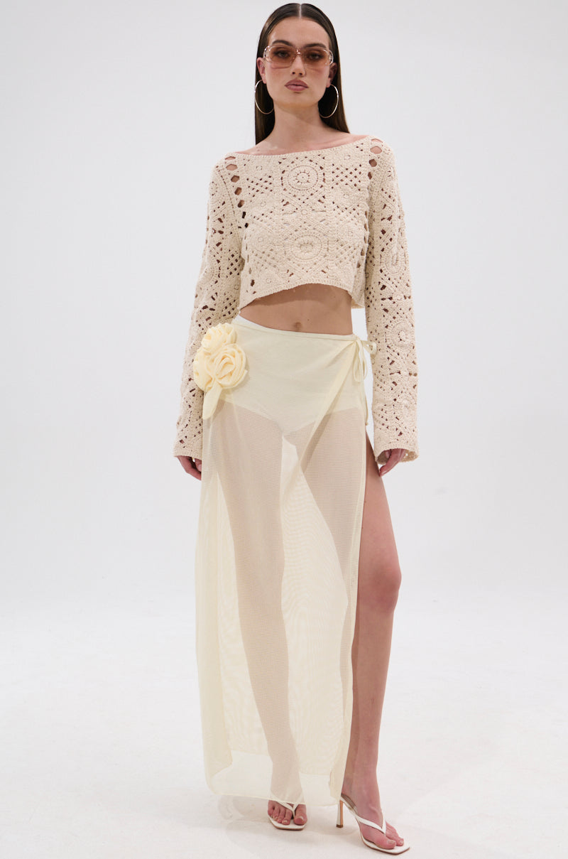 A woman stands against a plain white background wearing a cream crochet long-sleeve crop top, the CROCHET BEAUTY COVER UP SKIRT—a sheer pale yellow wrap skirt with a large flower accent—beige heels, hoop earrings, and tinted sunglasses. 5